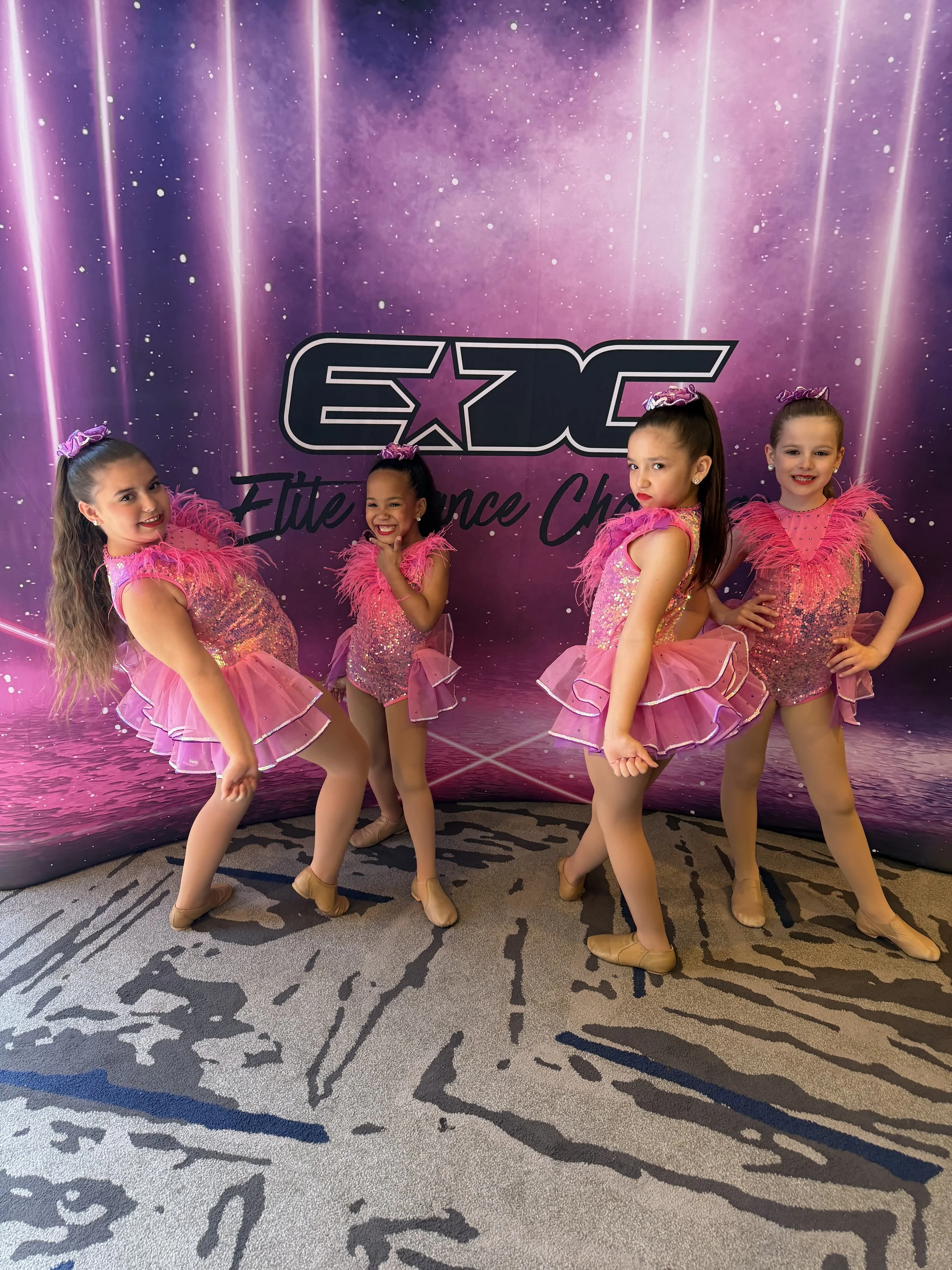 Four young girls in pink dance costumes pose in front of a purple backdrop with the words "EXC" and "Elite Dance Champs" written on it.