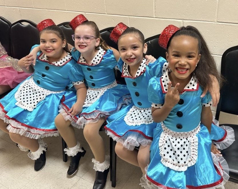 Four young girls dressed in colorful blue, white, and red polka dot costumes, sitting on chairs backstage, smiling and posing for the camera.
