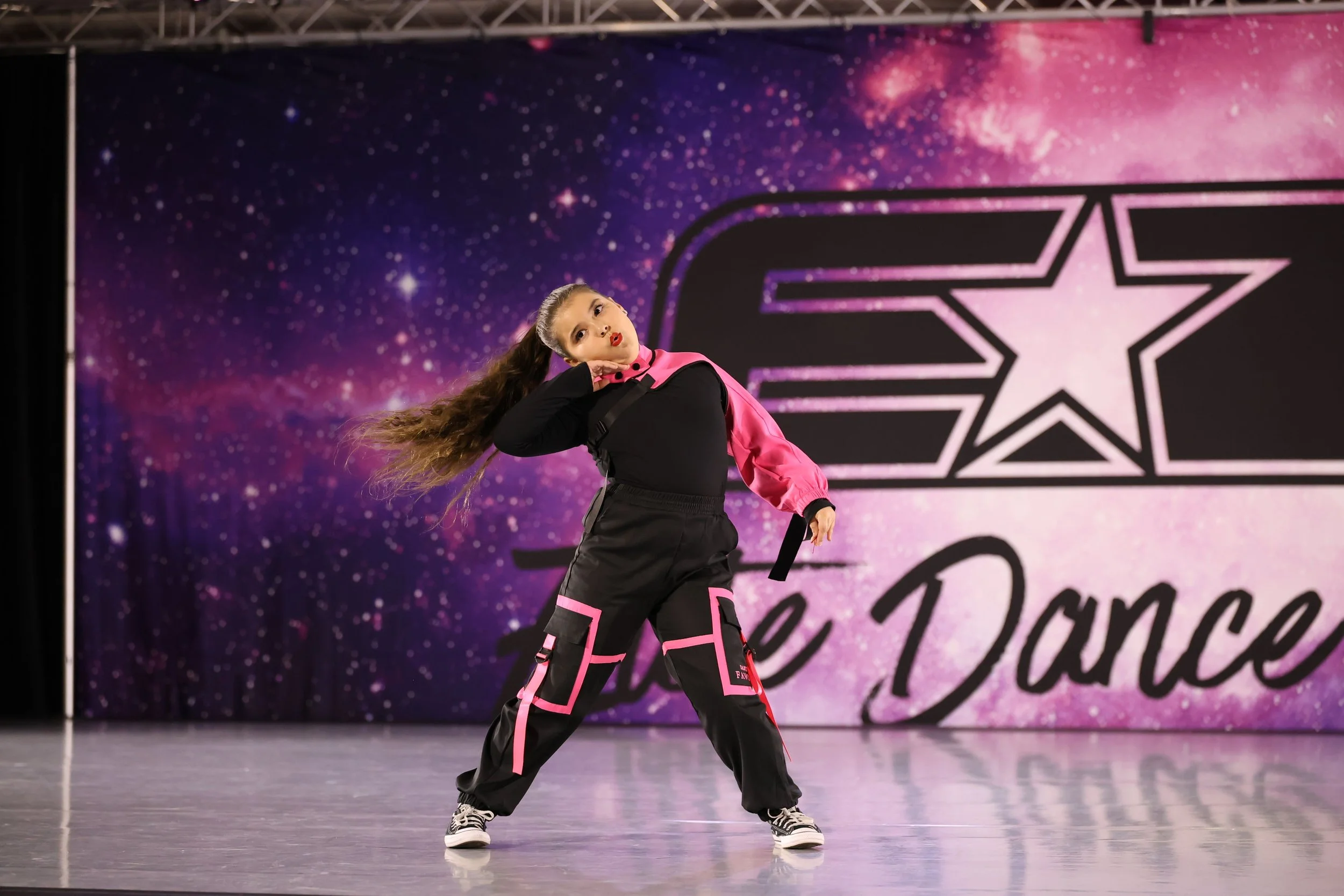 Young girl performing a dance routine on stage in front of a galaxy-themed backdrop with a pink star design, dressed in black and pink dance attire.