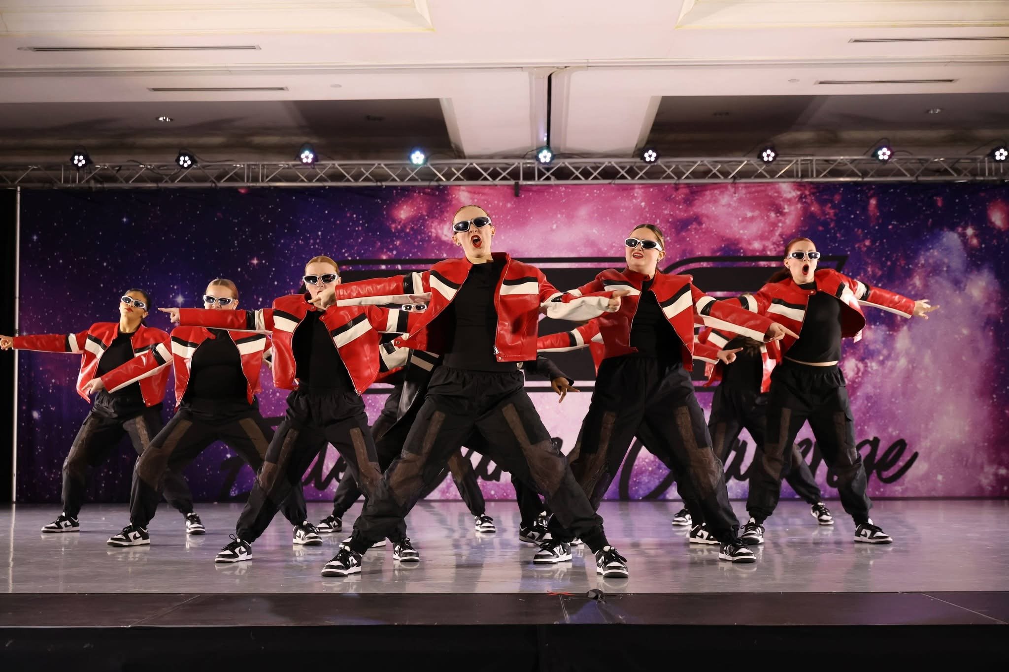 A group of dancers in red and black outfits performing on stage with a galaxy-themed backdrop.