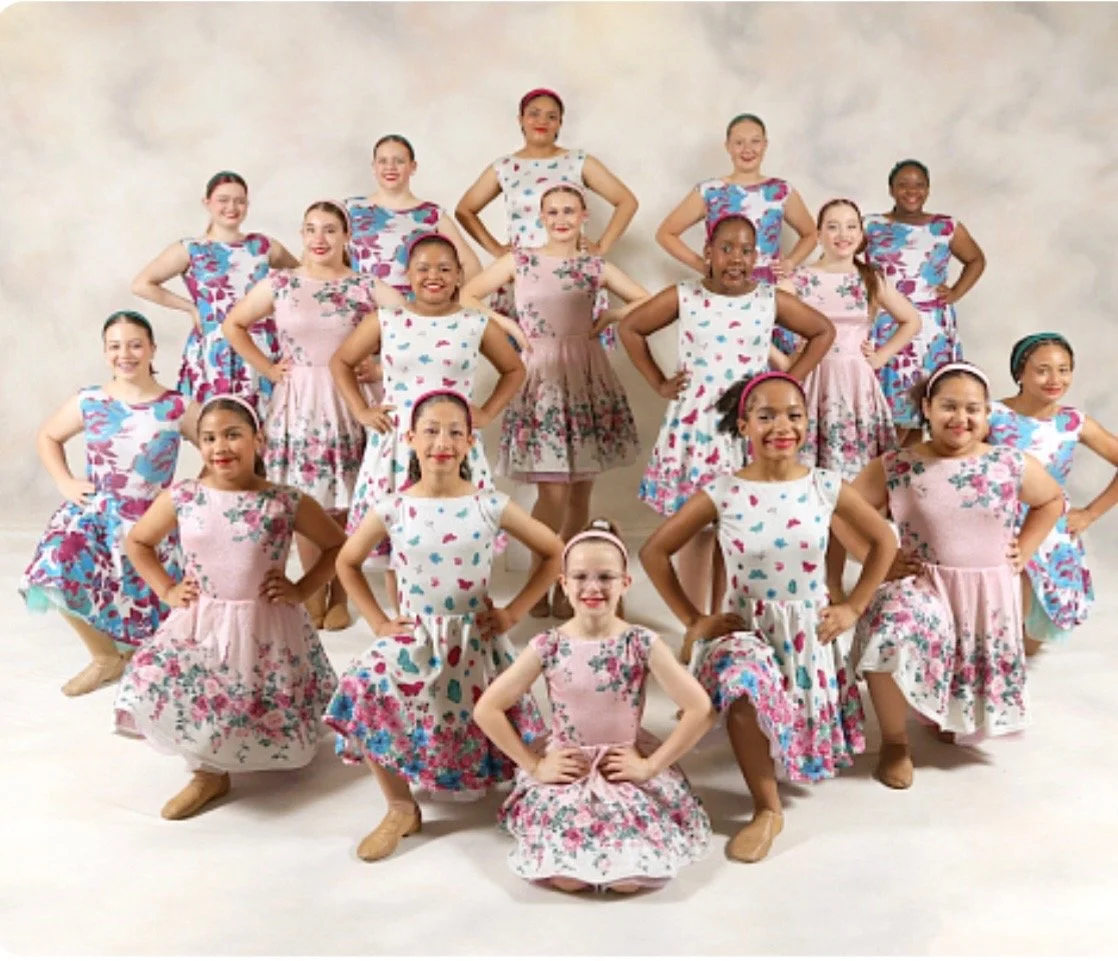 Group of young girls in coordinated floral dresses posing with hands on hips in a dance studio.