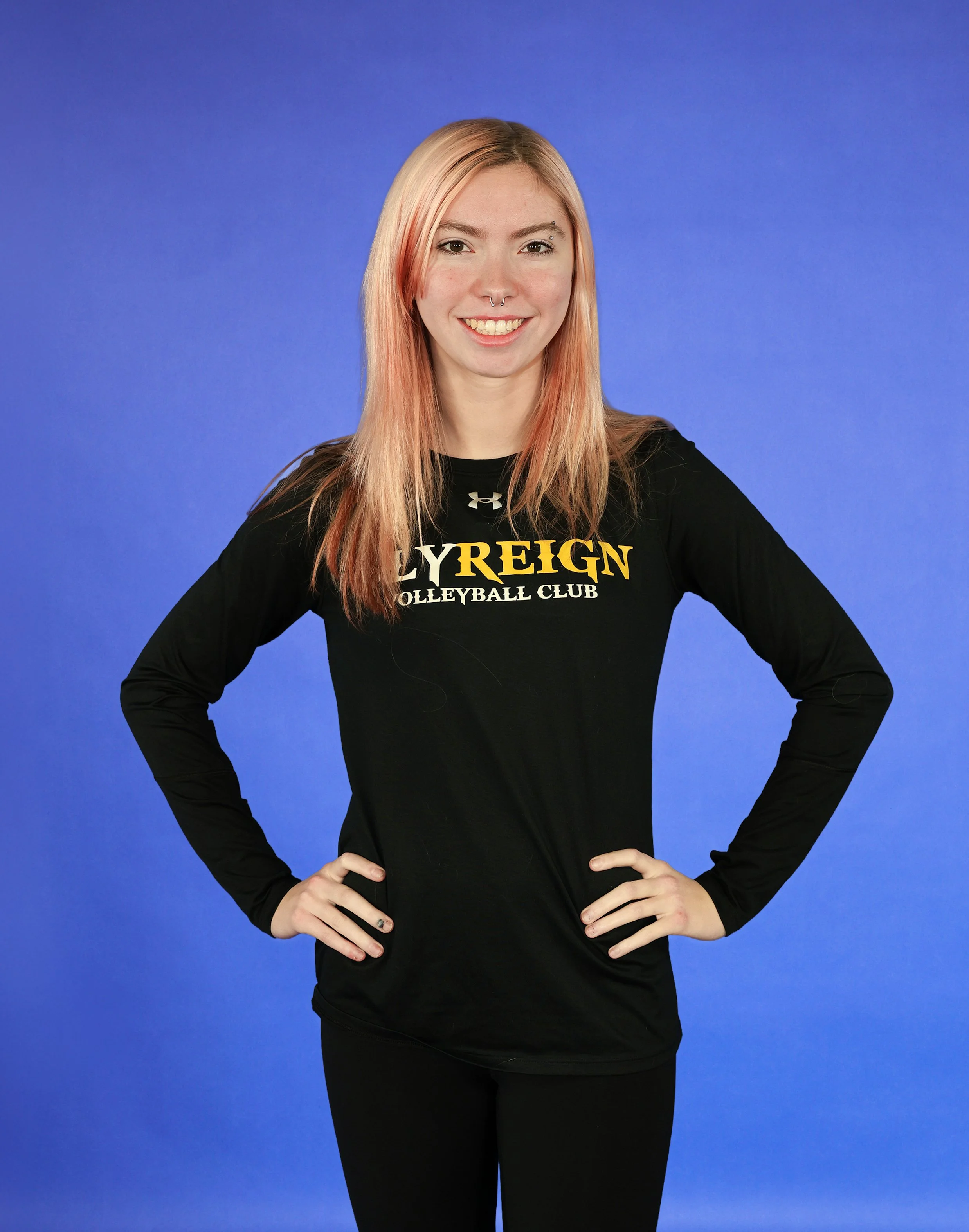 A woman with long pink hair, wearing a black long-sleeve shirt with yellow and white lettering, poses with her hands on her hips against a blue gradient background.