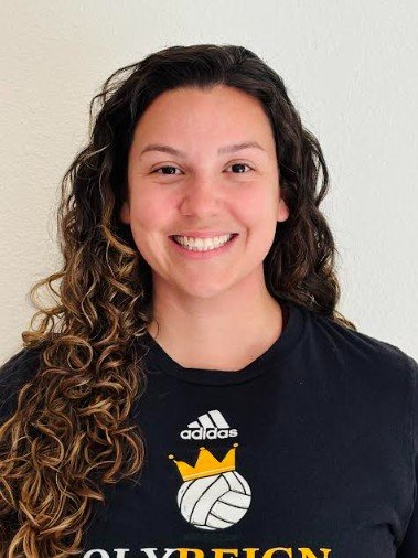 Young woman with curly hair smiling, wearing a black sports jersey with a volleyball graphic and a crown, standing against a plain off-white wall.