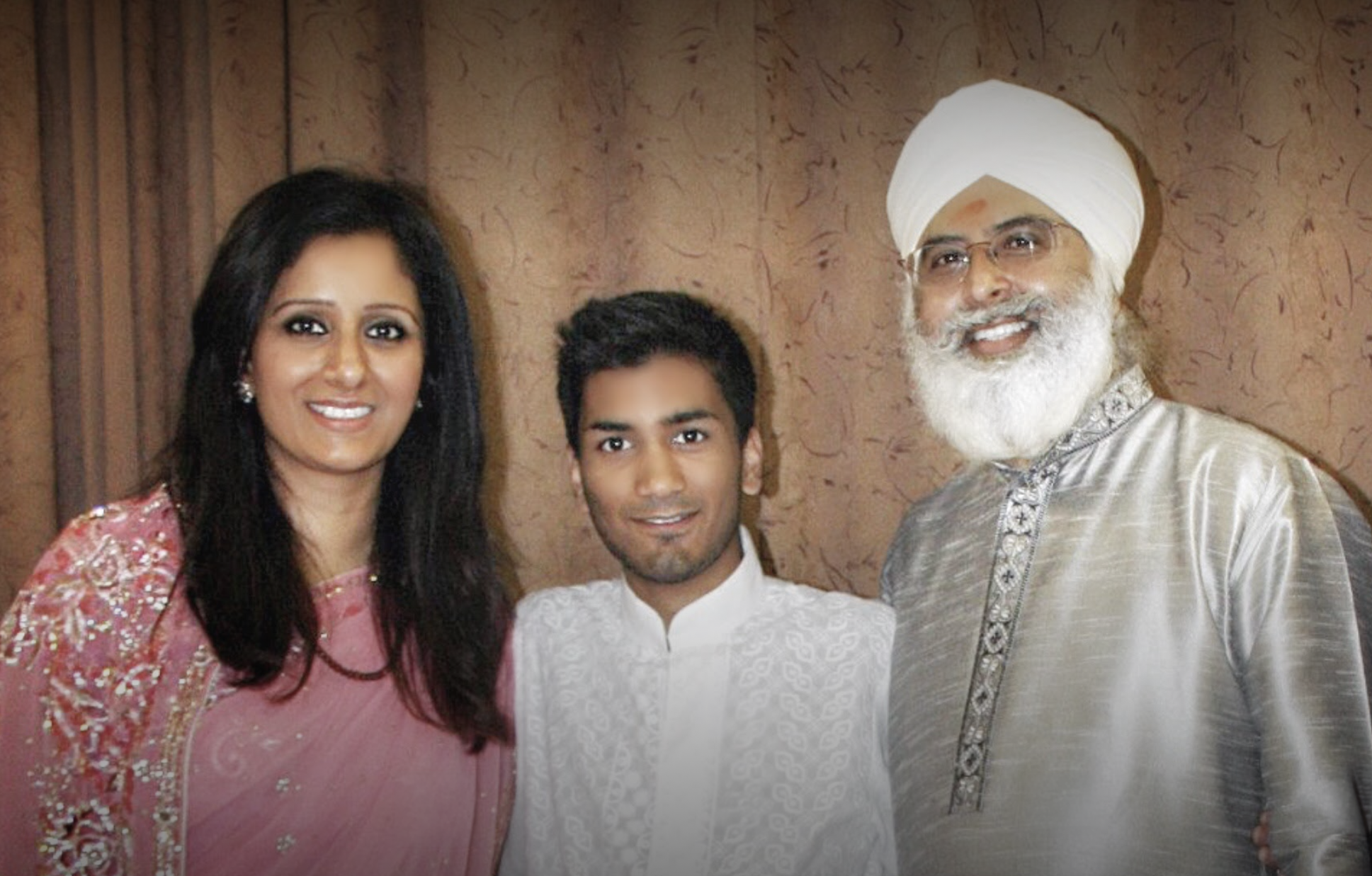 A family of four, dressed in traditional Indian attire, standing together indoors, smiling at the camera. The woman is wearing a pink and gold dress, the man a cream-colored kurta with embroidery, and an elderly man in a white turban and gray kurta.
