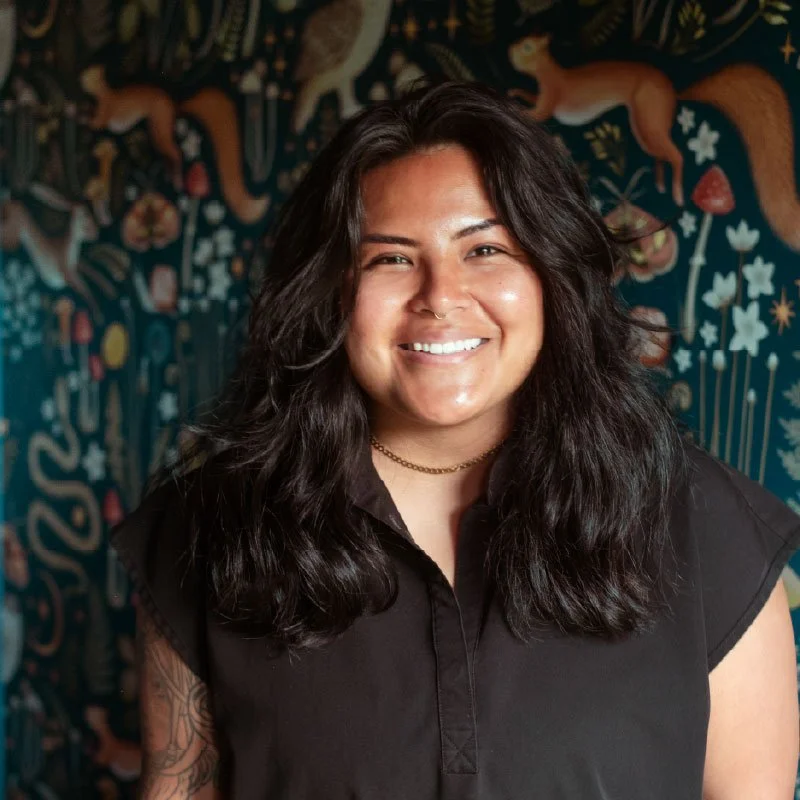 Smiling woman with long dark hair standing in front of a colorful mural featuring various animals and plants.