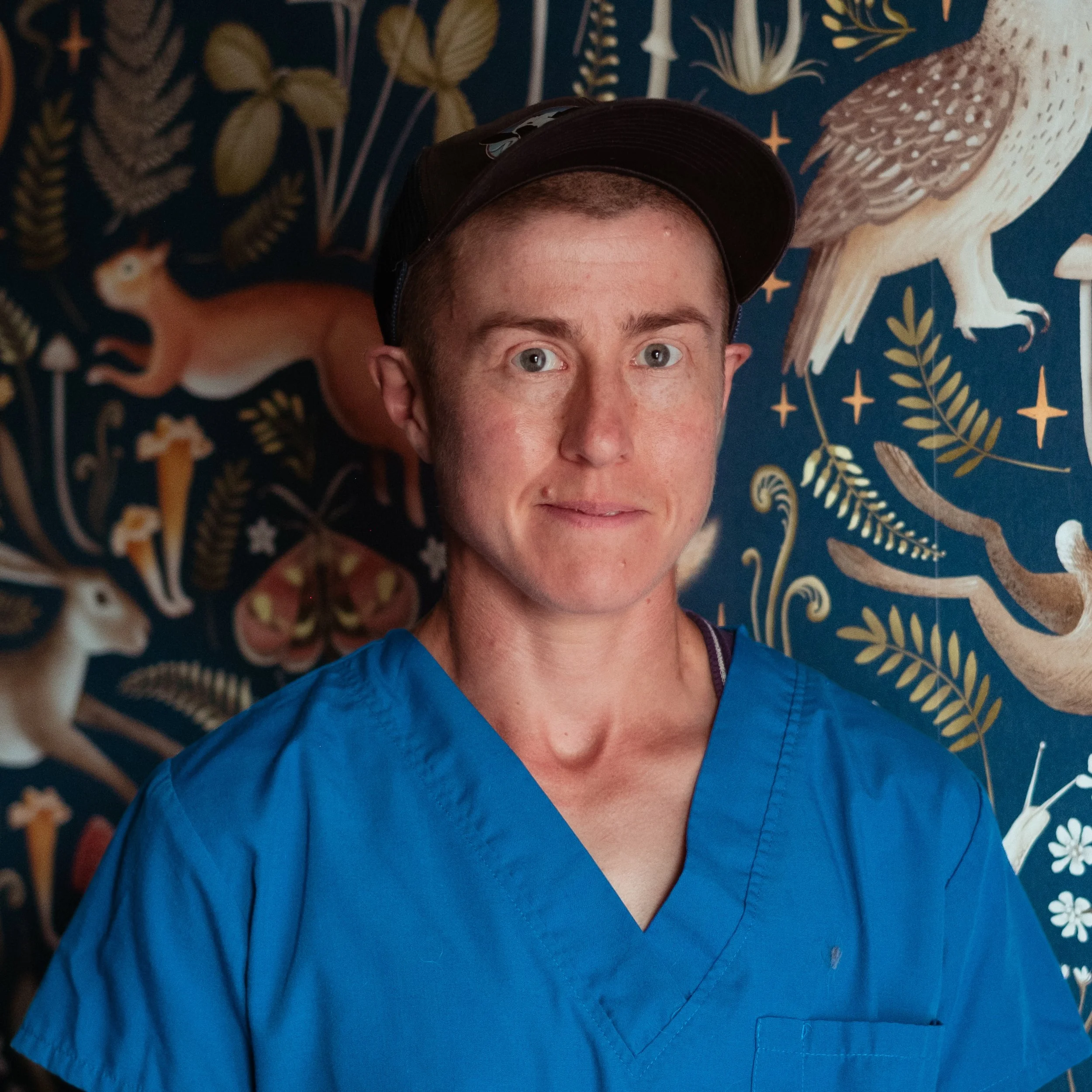 Close-up of a person with short brown hair and light skin, wearing a black cap and blue scrubs, standing in front of a decorative wallpaper with illustrations of animals and plants.