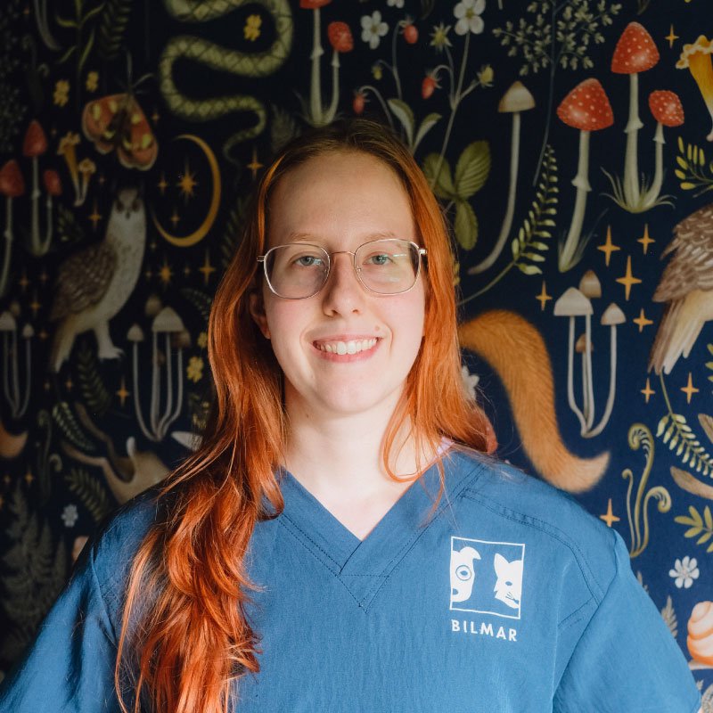 Young woman with long red hair and glasses smiling in front of a dark, whimsical forest-themed mural featuring various animals, mushrooms, and plants. She is wearing a blue medical scrub top with a logo that says "BILMAR."