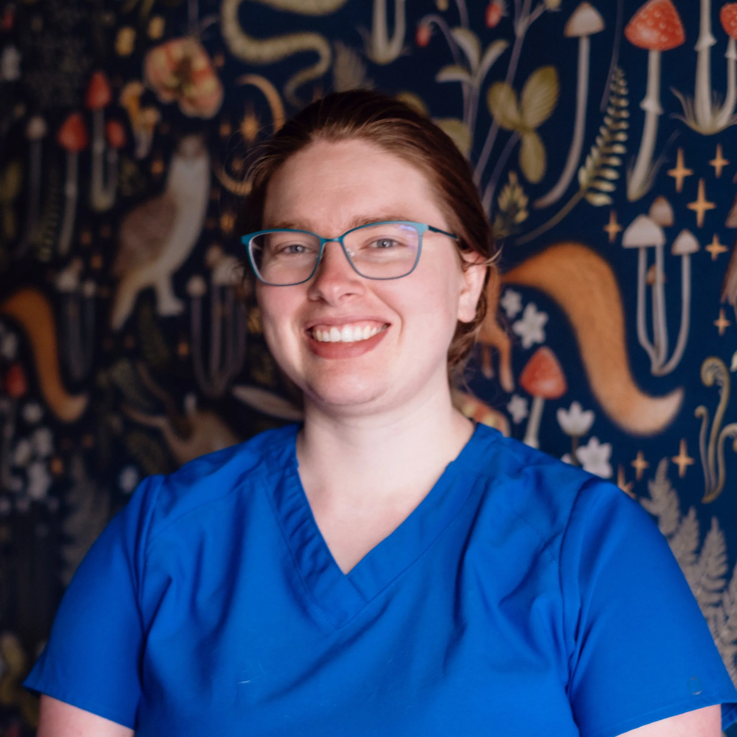 A person with red hair and glasses wearing a blue shirt smiling for a selfie against a dark background with a gold and white ornate pattern.