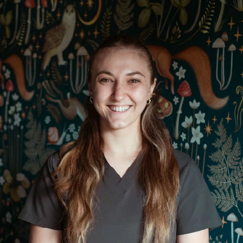 Smiling woman with long brown hair and earrings standing in front of a dark, patterned background with animals and plants.