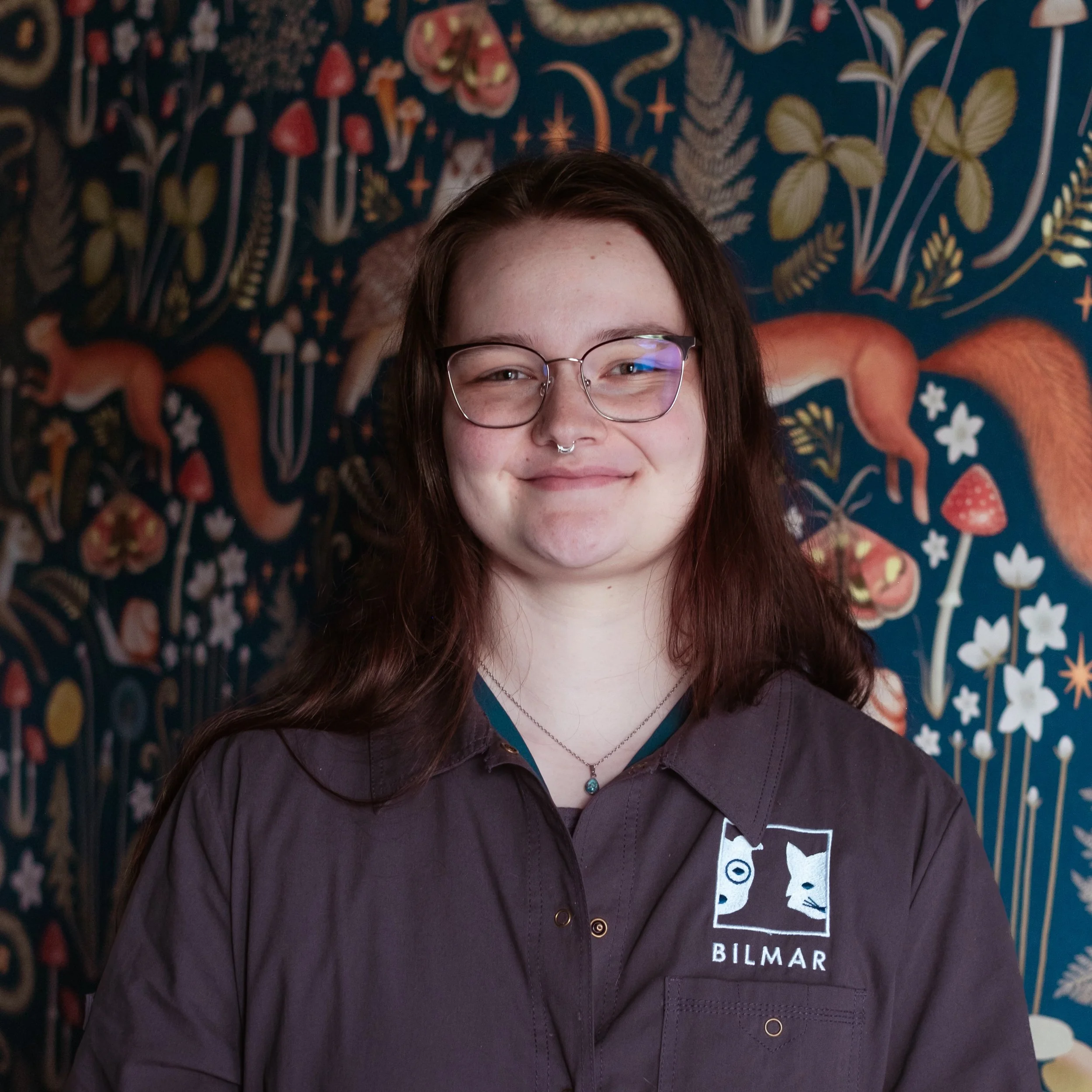 Close-up of a person with long brown hair, wearing glasses, a septum piercing, smiling, standing in front of a colorful botanical-themed background with illustrations of plants, flowers, and fungi. The person is wearing a dark-colored shirt and a necklace with a blue pendant.