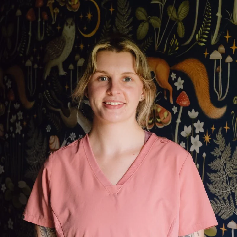 A woman with short blond hair wearing a pink scrub top standing in front of a dark botanical and animal-themed mural.