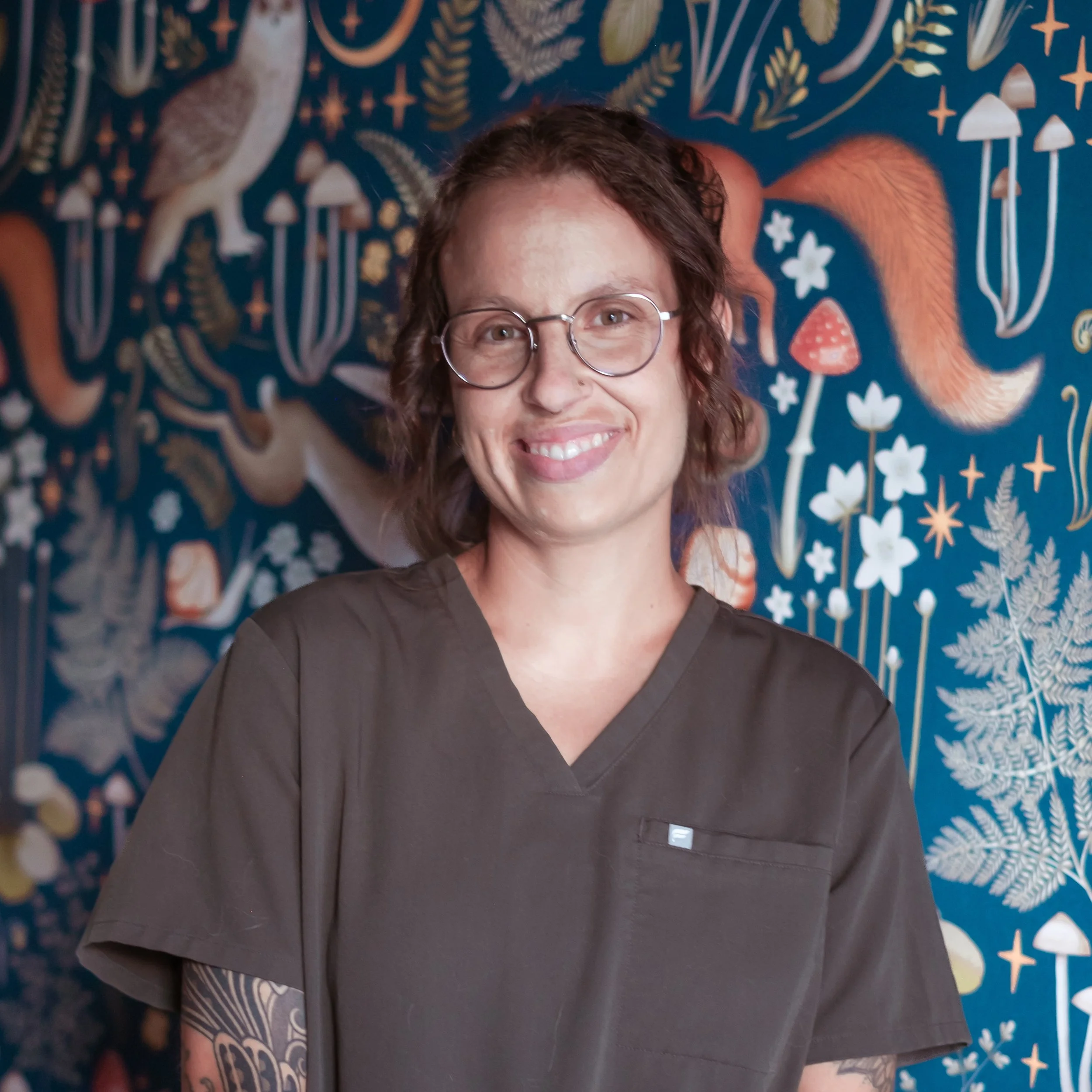 A woman with curly brown hair and glasses smiling in front of a colorful mural featuring mythical creatures, flowers, stars, and abstract designs.