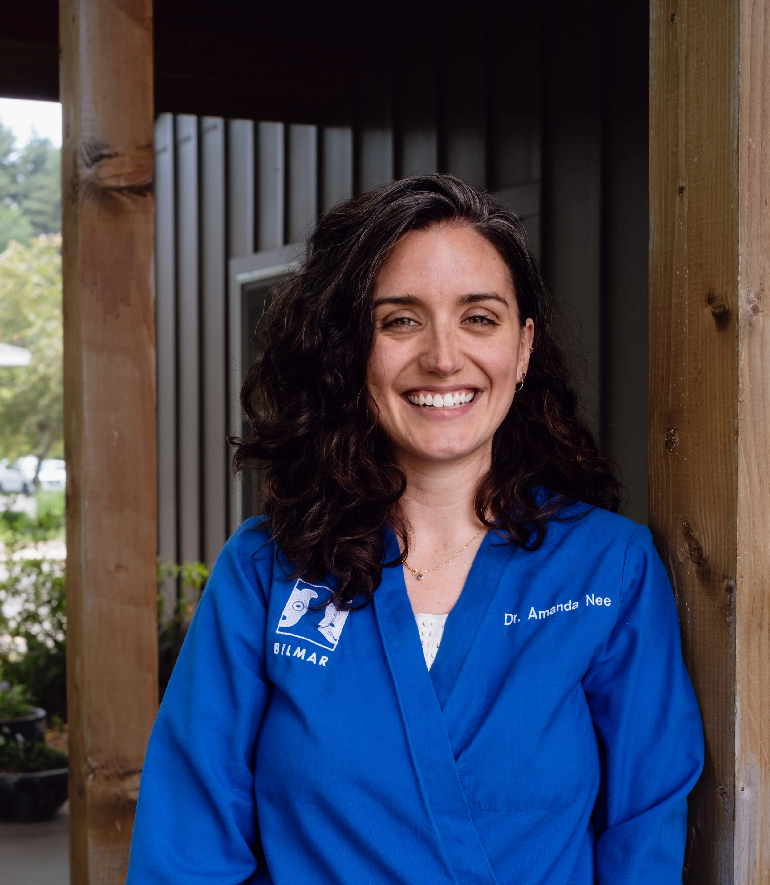 Close-up of a woman with dark, curly hair smiling, wearing a blue medical jacket with text. She is standing outdoors among greenery, framed by wooden beams.