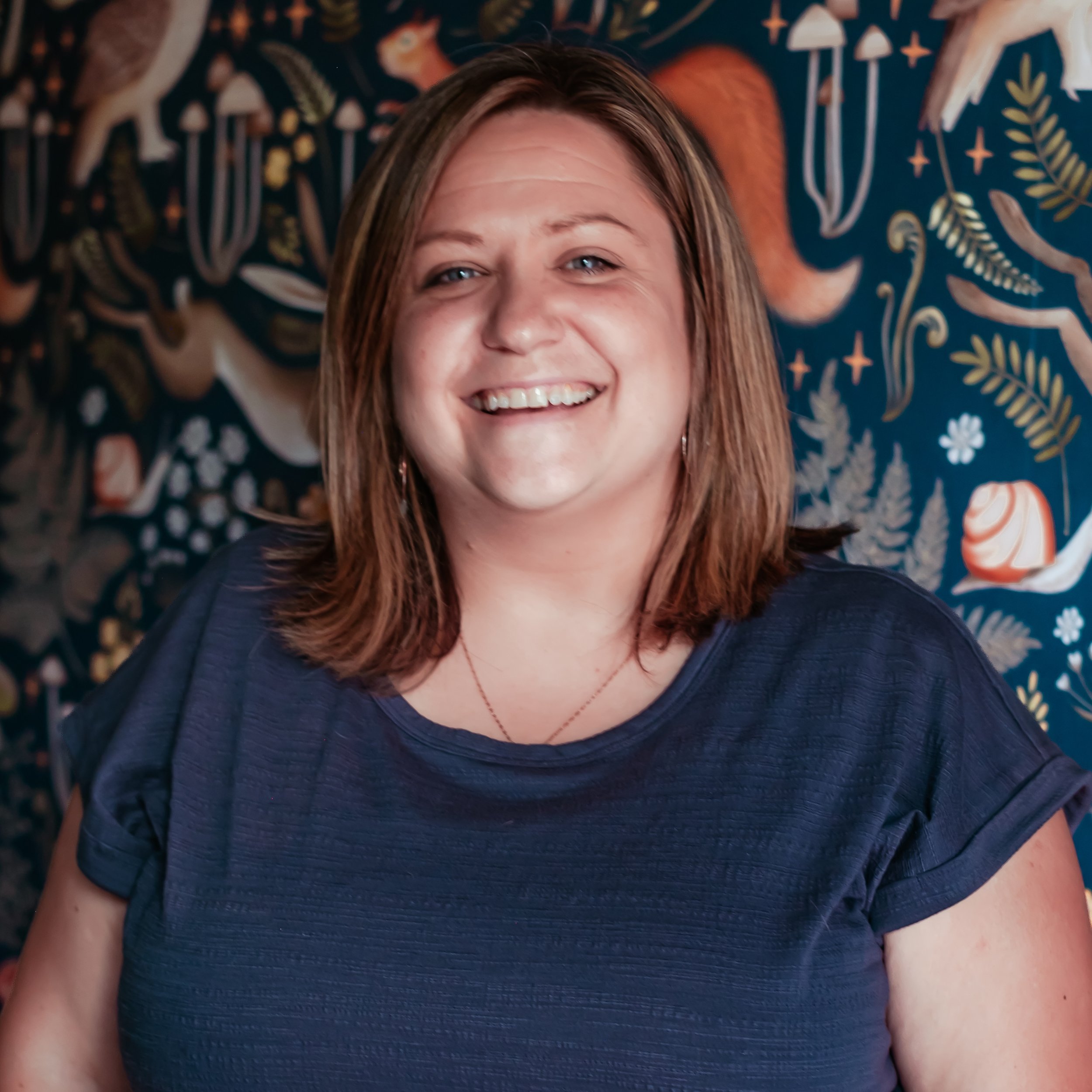 Close-up of a smiling woman with light brown hair wearing a navy blue top, standing in front of a colorful, patterned tapestry with abstract shapes and animals.