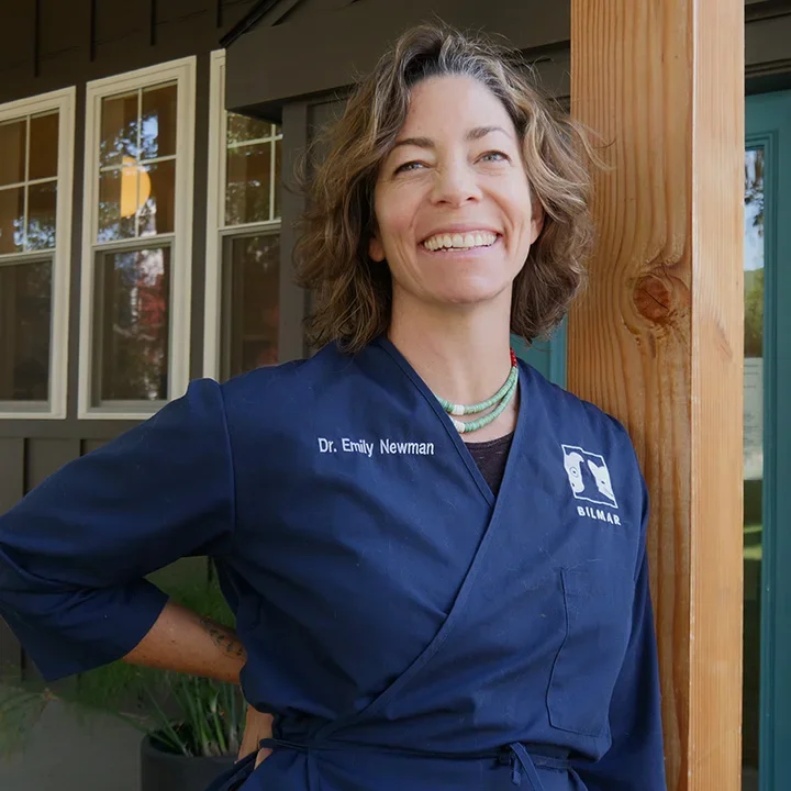 A woman smiling and standing outdoors in front of a house with large windows, wearing a blue chef's coat with