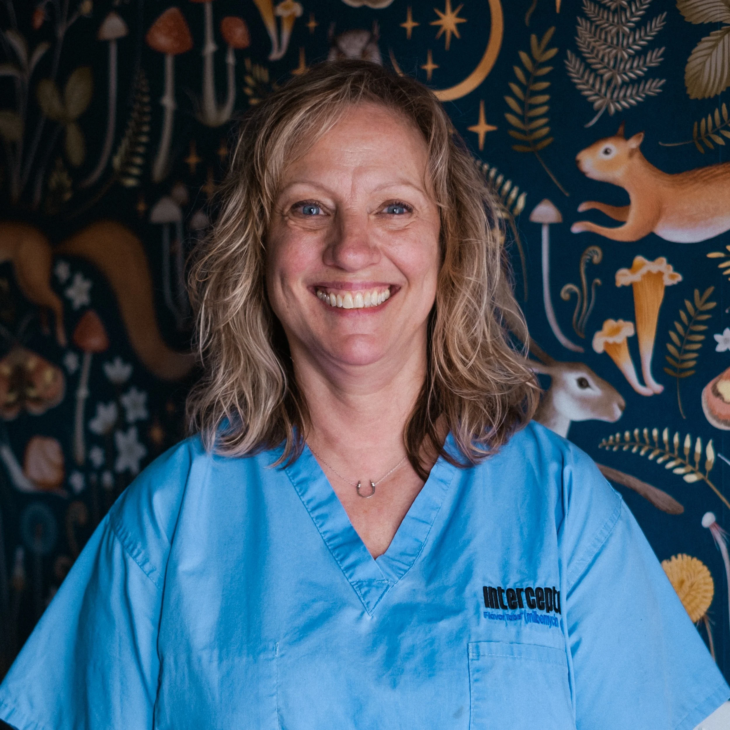 Close-up of a smiling woman wearing blue scrubs with the logo 'mercy' on it, standing in front of a dark blue wall with a celestial and animal-themed pattern.