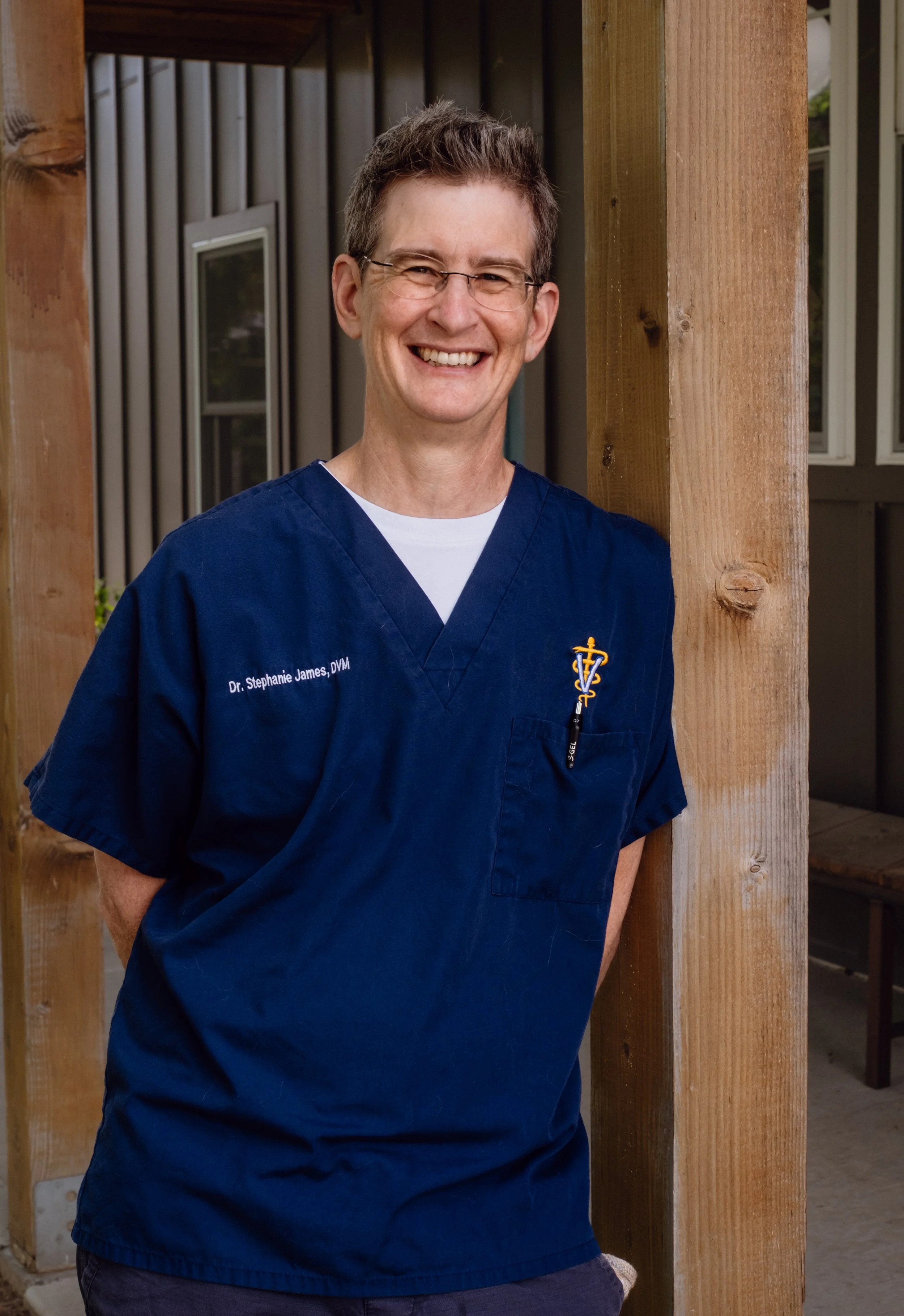 A smiling woman with short brown hair and glasses wearing a navy blue veterinary uniform standing outdoors next to a wooden post.