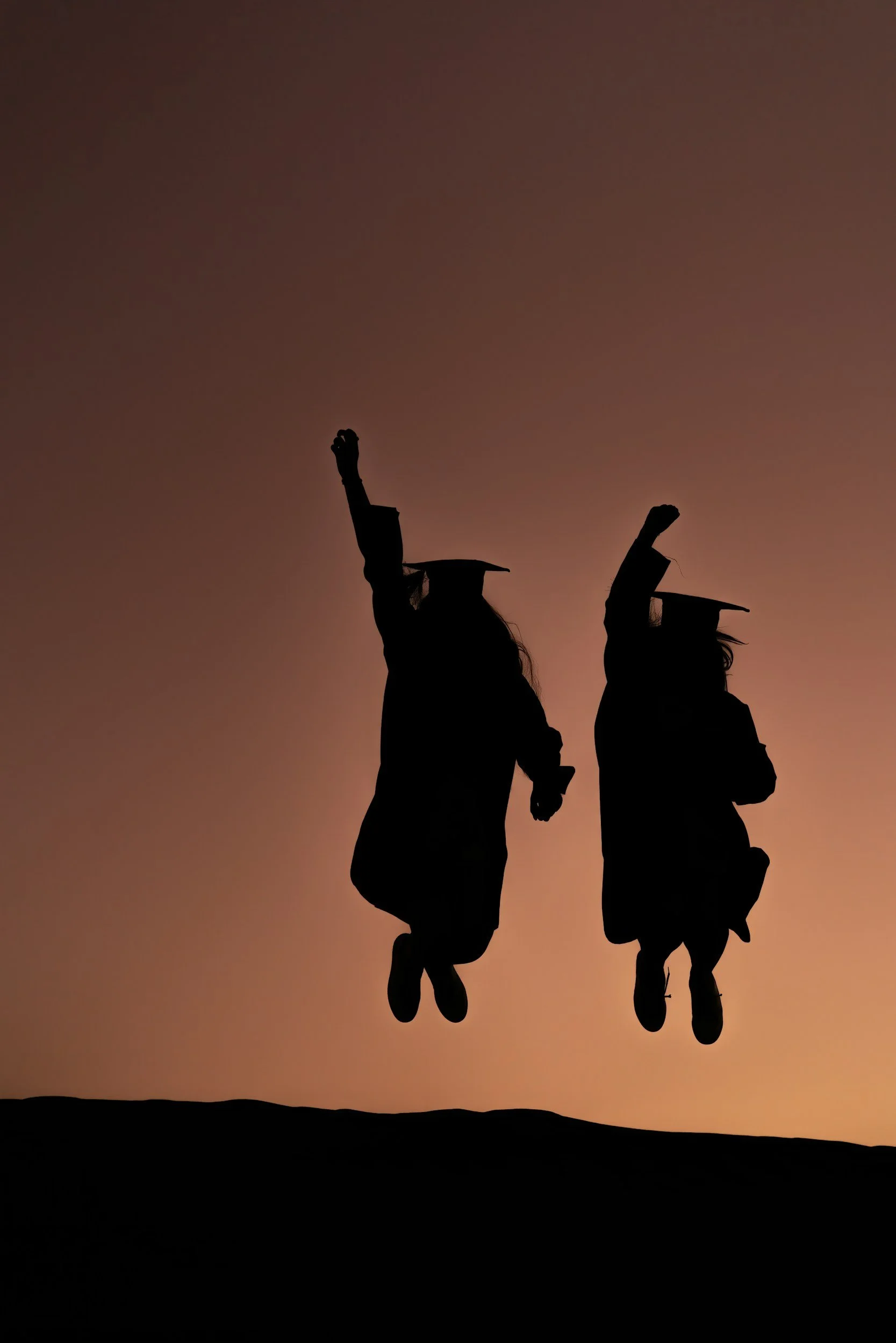 The silhouette of two women in graduation robes and caps jumping in front of a subset.