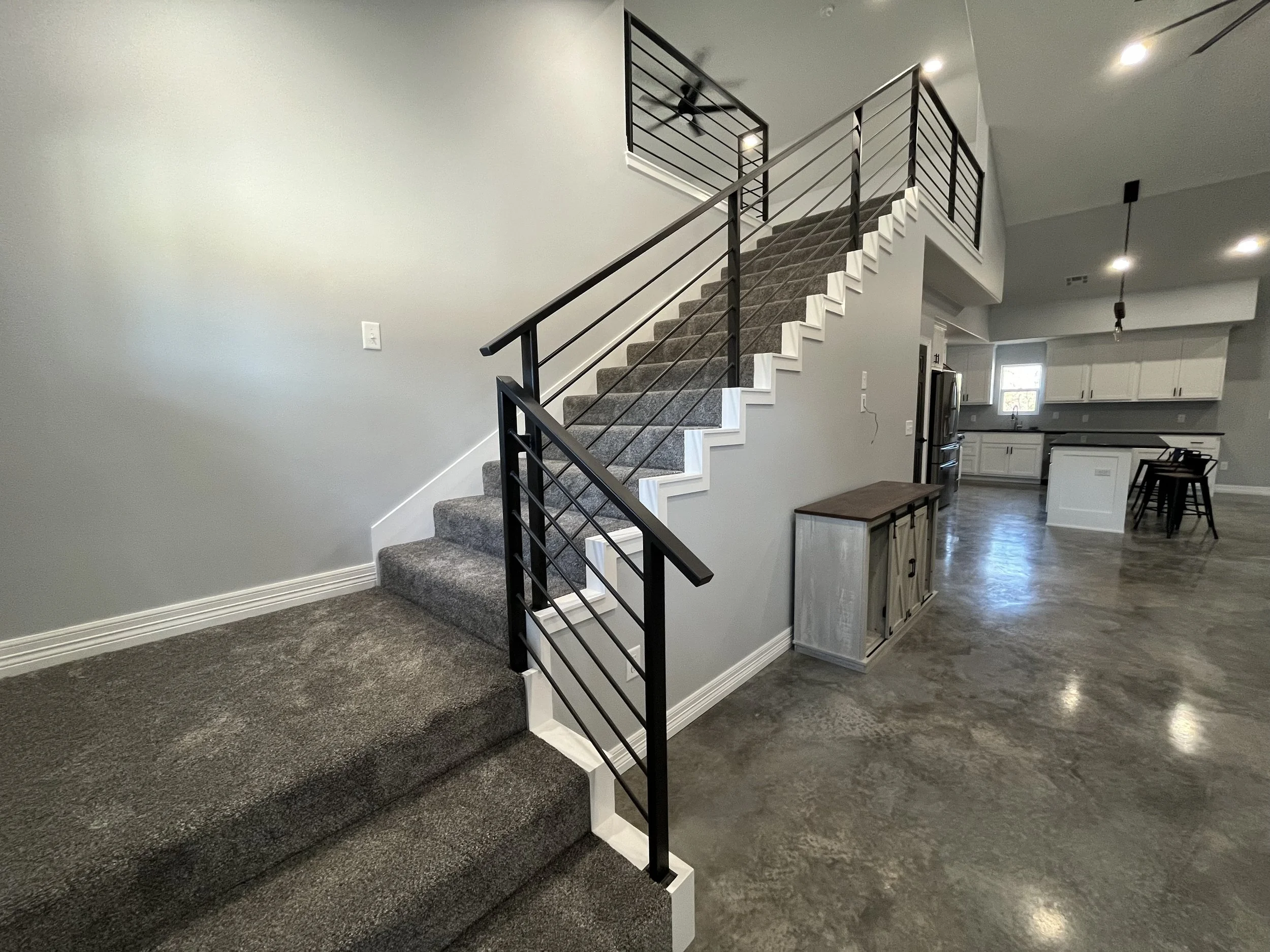 Interior of a modern open-concept home featuring a carpeted staircase with black railings leading to an upper floor, a large living space with gray concrete floors, and a kitchen with white cabinets, stainless steel appliances, and a dining table with chairs.