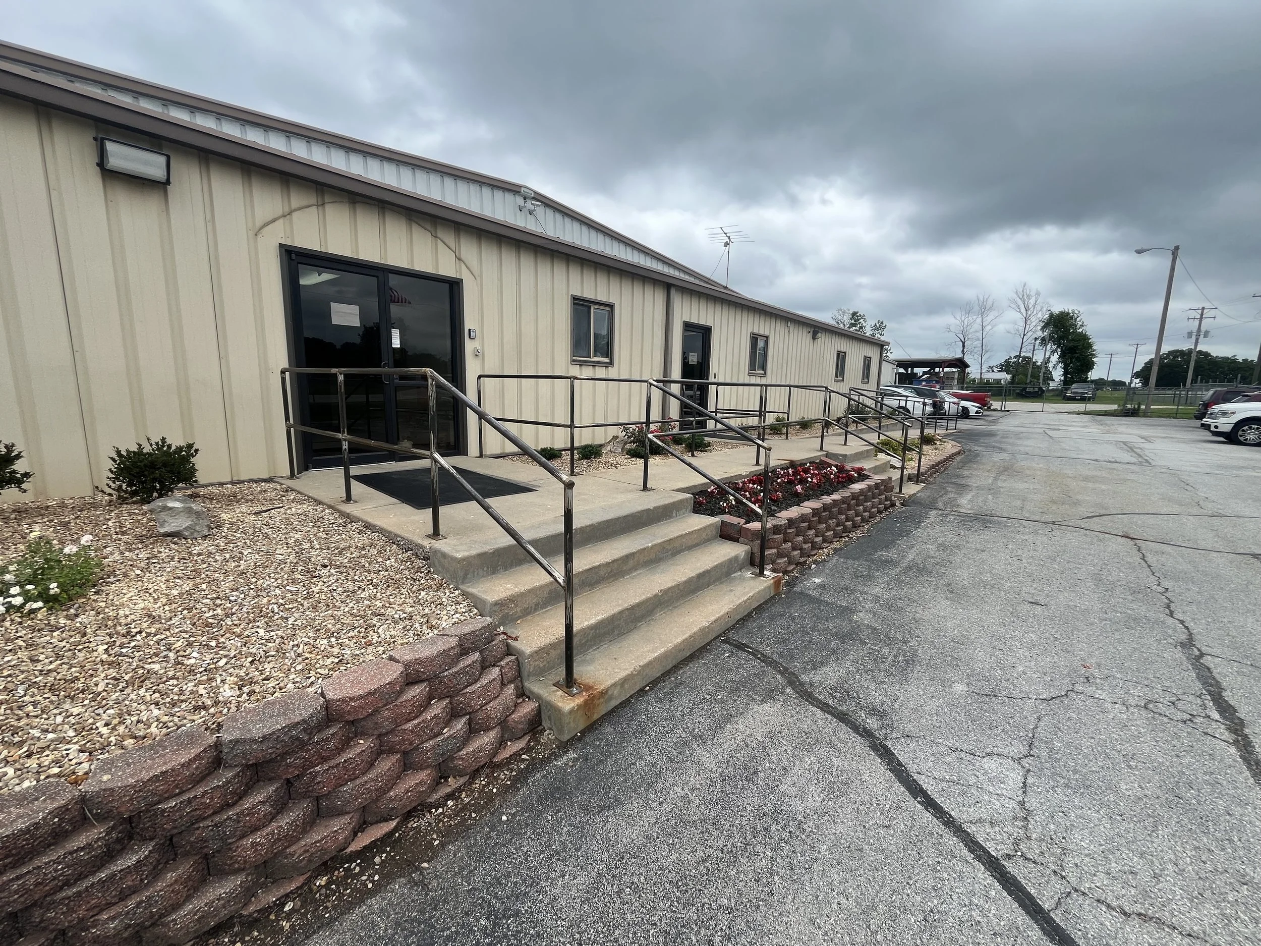 Exterior view of a beige commercial building with entrance accessed by concrete stairs and a wheelchair ramp, surrounded by Commercial handrail and parking lot with some cars, under an overcast sky.