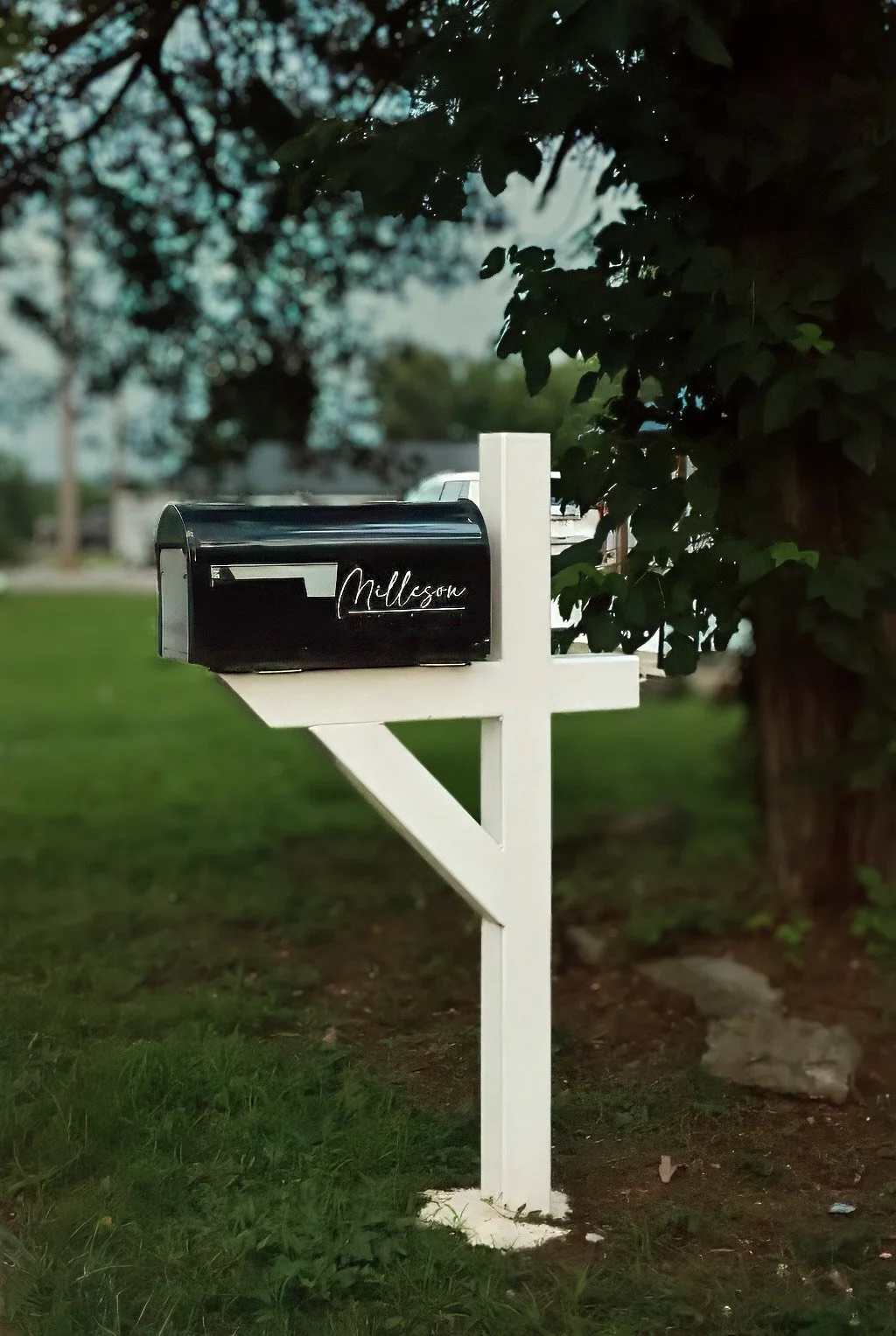 Black metal mailbox with white script that says 'Milson' mounted on a white wooden post next to a large leafy tree, in a yard with green grass.