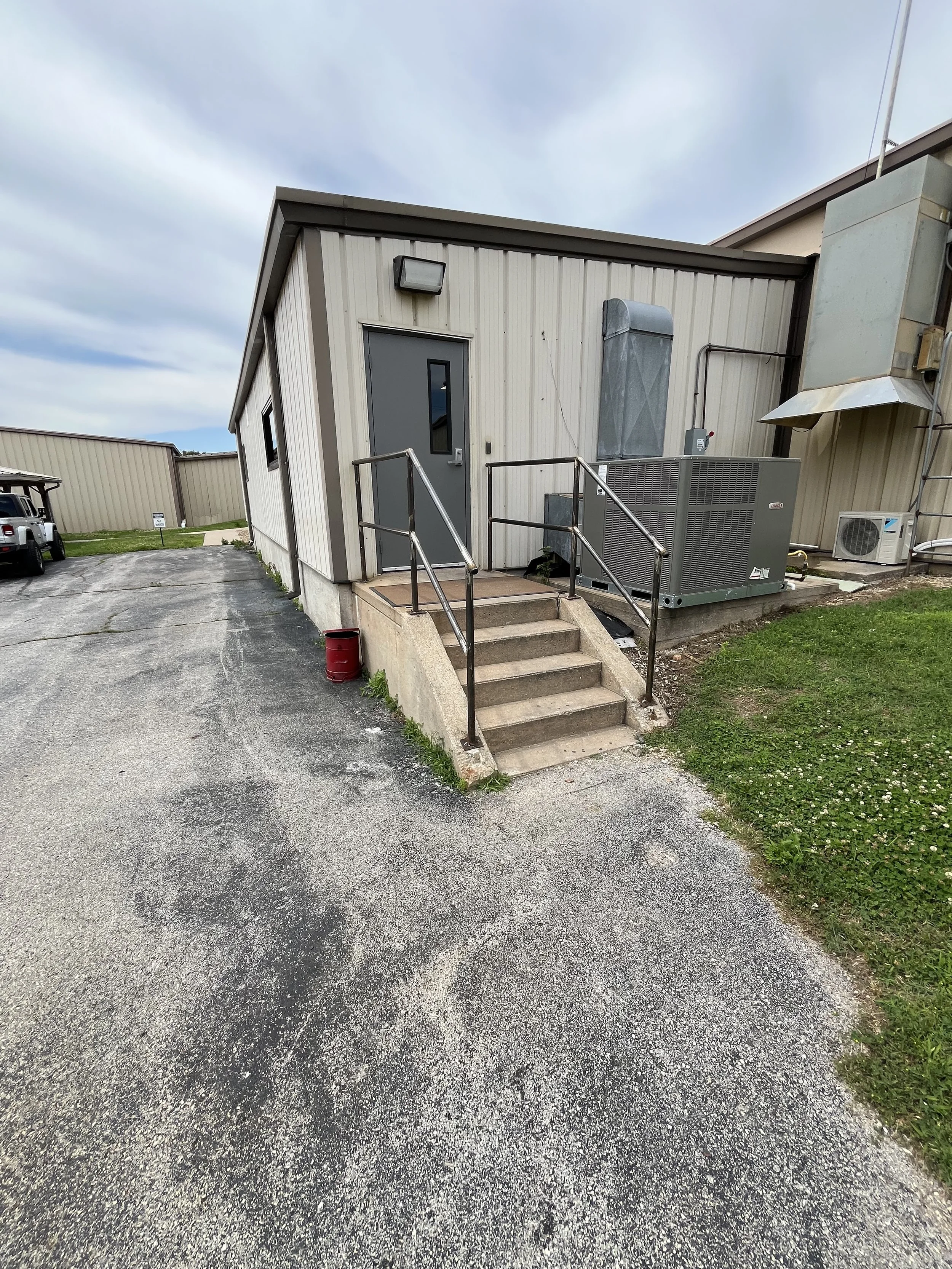 Exterior of a beige metal building with small staircase and metal railing leading to a gray door. There is a grassy area and parking lot nearby.