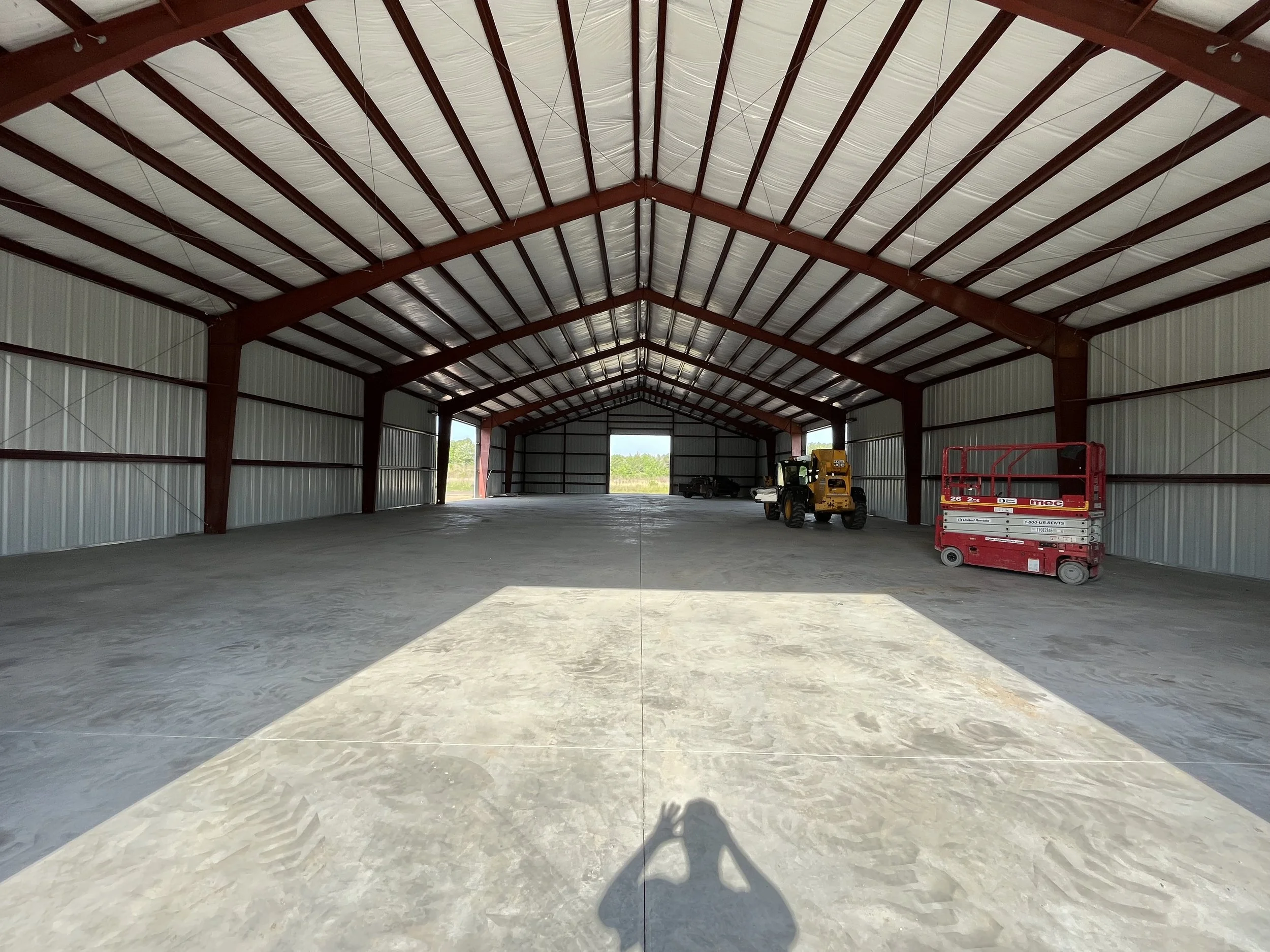 Inside an unfinished large metal warehouse with a concrete floor, a mini forklift, and a red scissor lift, with sunlight coming through open garage doors.