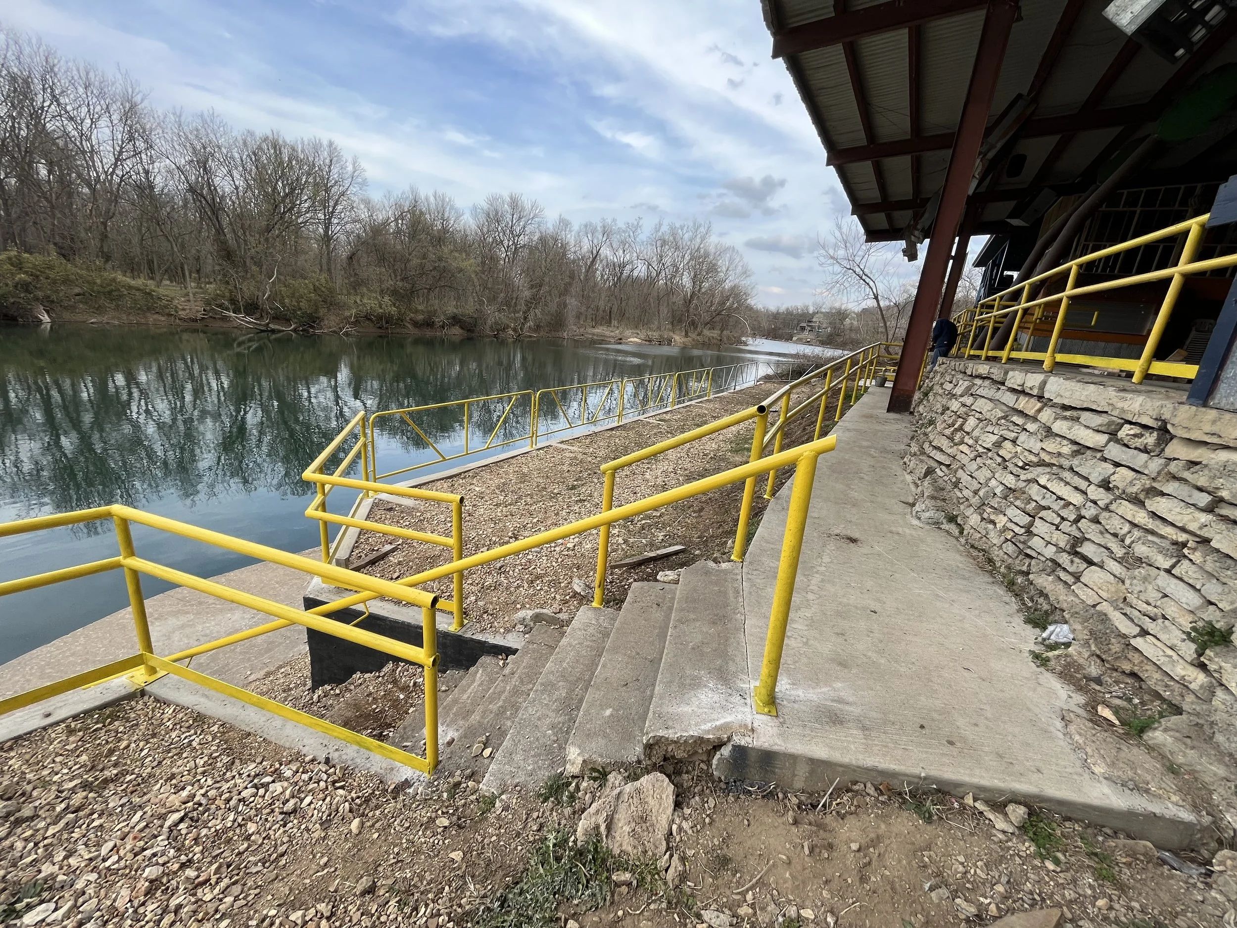 View of a riverside walkway with yellow metal railings, concrete stairs, and a stone wall, overlooking a calm river surrounded by leafless trees under a partly cloudy sky.