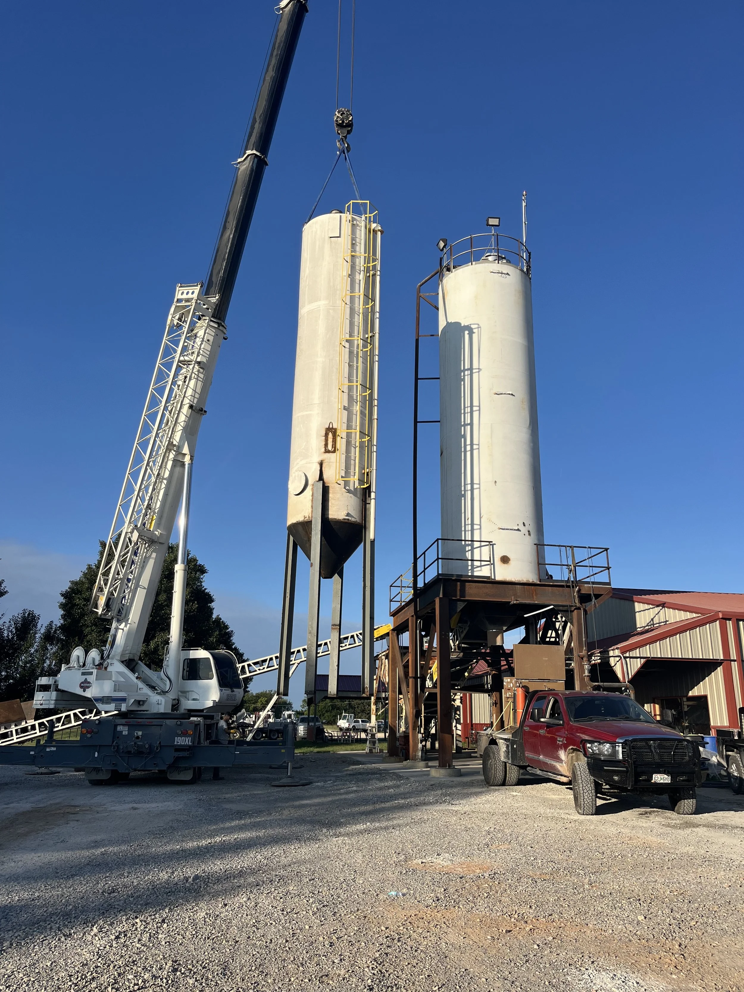 Construction site with a crane lifting a large metal storage tank next to two other tall tanks, with a red pickup truck parked nearby and a building in the background.