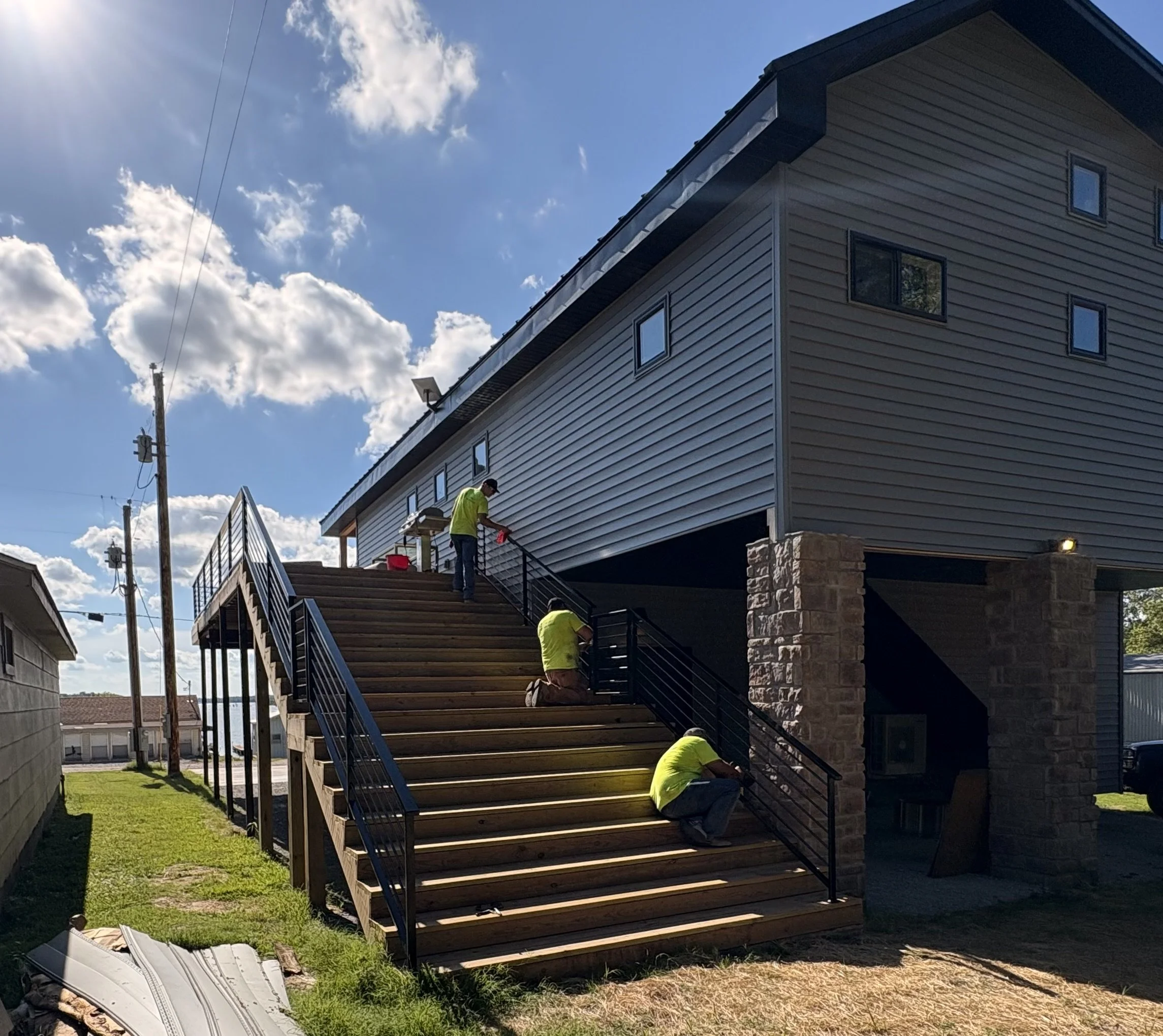 Three workers in yellow shirts installing a new wooden staircase with black metal railings leading up to a house with gray siding and a brick foundation under a sunny sky.