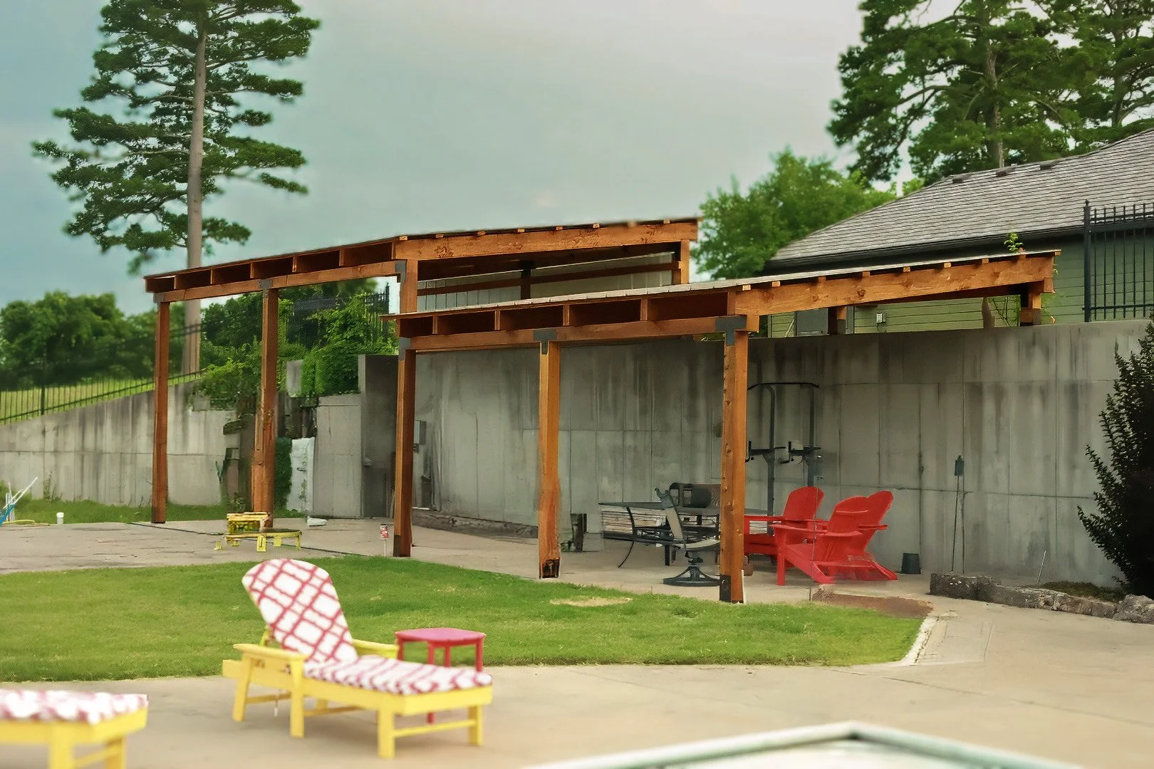 Backyard patio area with a concrete wall, outdoor furniture, including red chairs, and an unfinished wooden pergola structure. There are trees and a house in the background.
