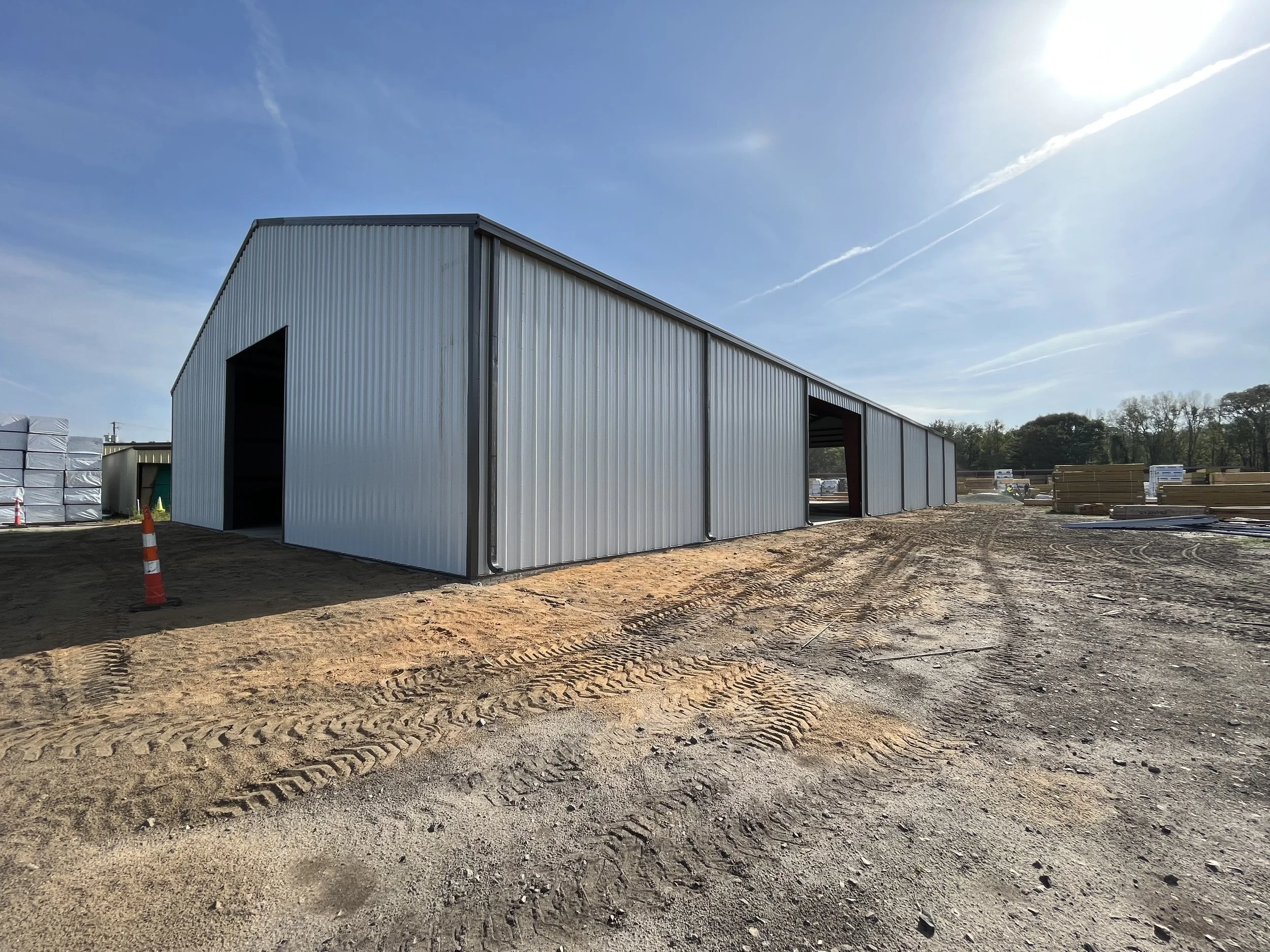 Construction site with a newly built metal storage or workshop building, dirt ground with tire tracks, clear sky, and some construction materials and equipment in the background.