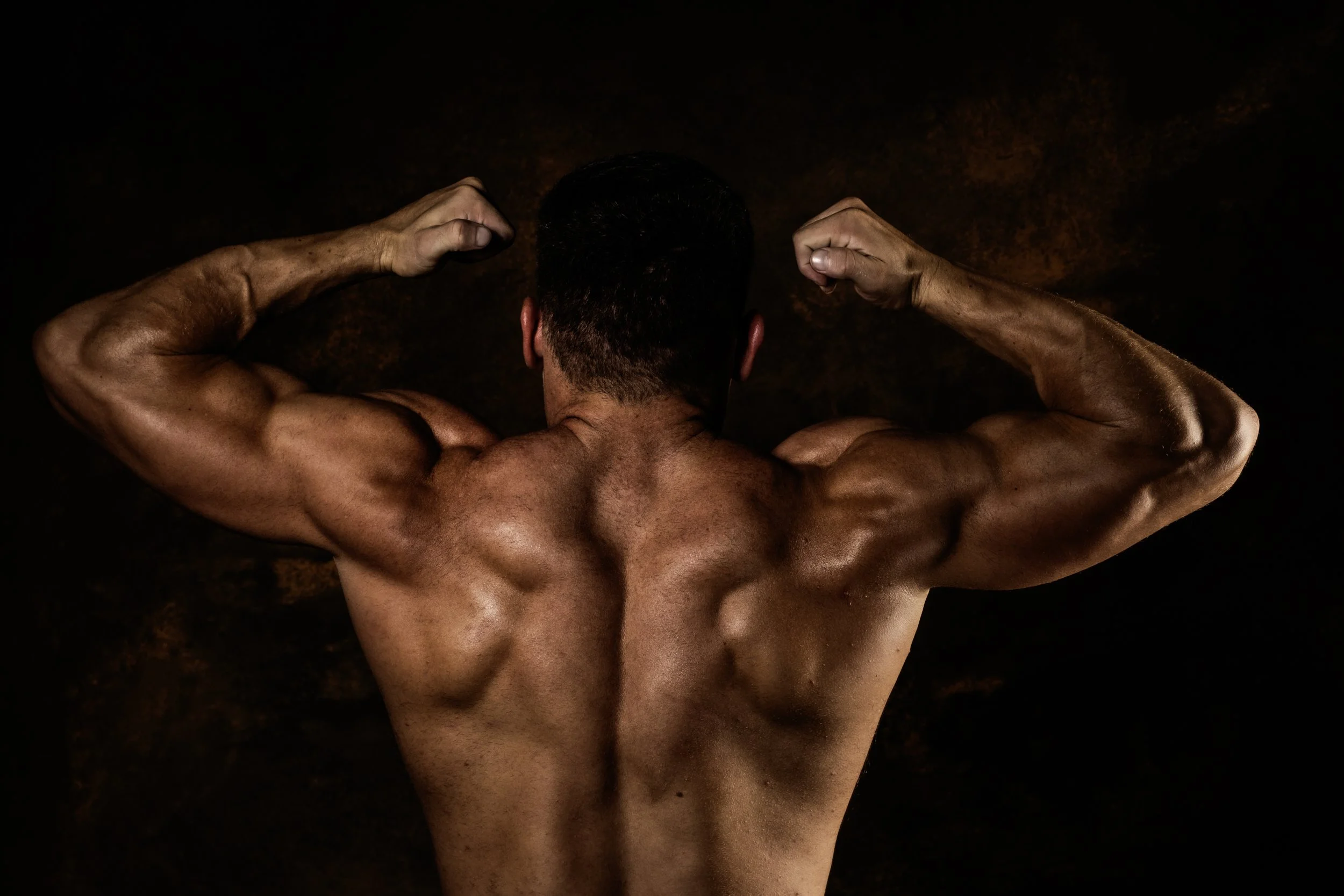 Back view of a muscular man flexing his biceps with his arms raised, dark background.