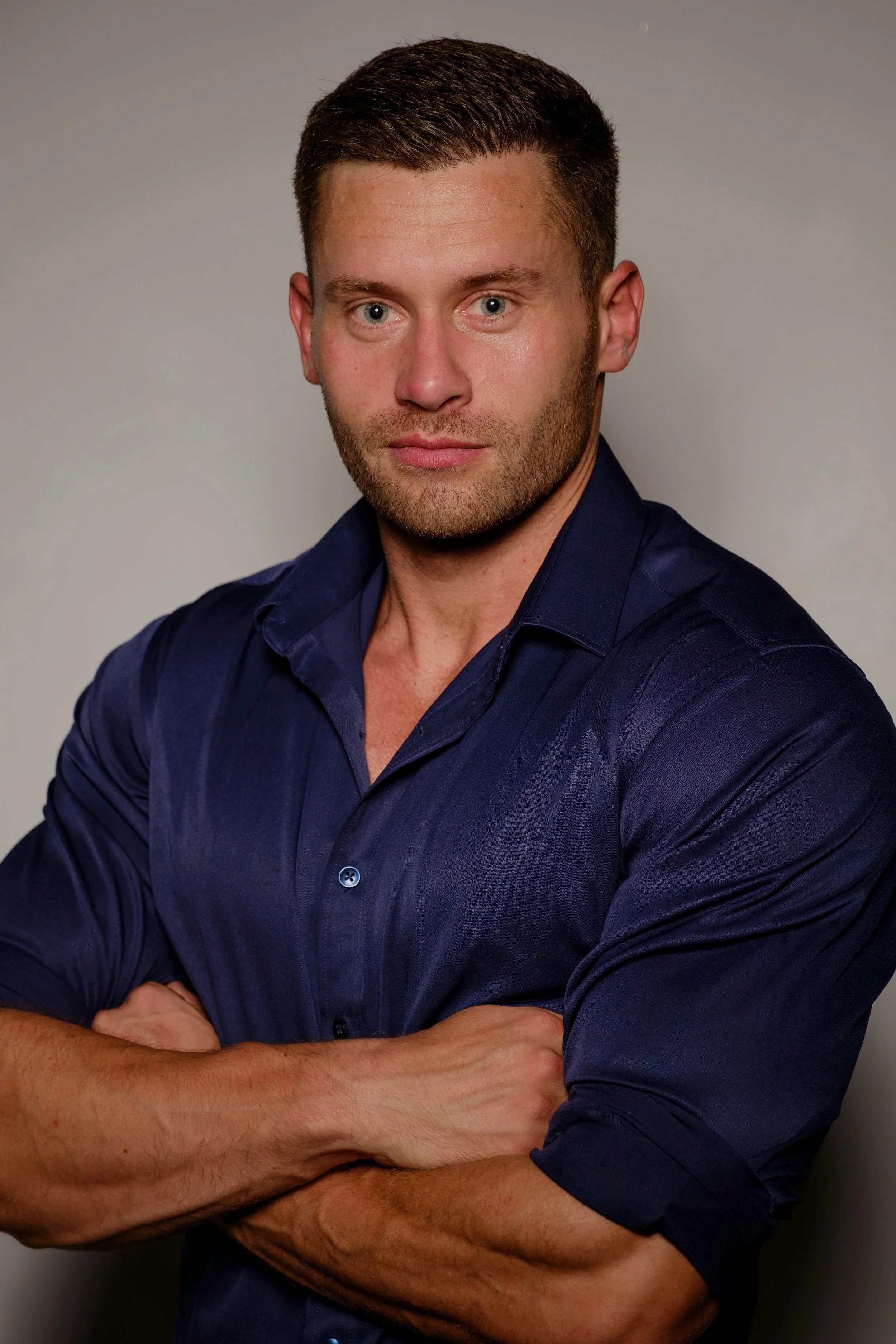 A young man with short brown hair, light stubble, and blue eyes, wearing a navy blue button-up shirt with sleeves rolled up, standing against a plain light-colored background.