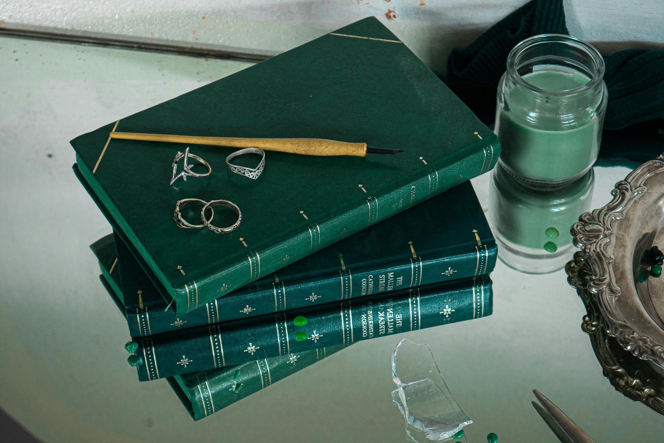 A mirror with a broken glass piece, three hardcover green books stacked with rings and a jewelry piece on top, a jar of green nail polish, a decorative metallic tray, a candle, and a pair of tweezers on a reflective surface.