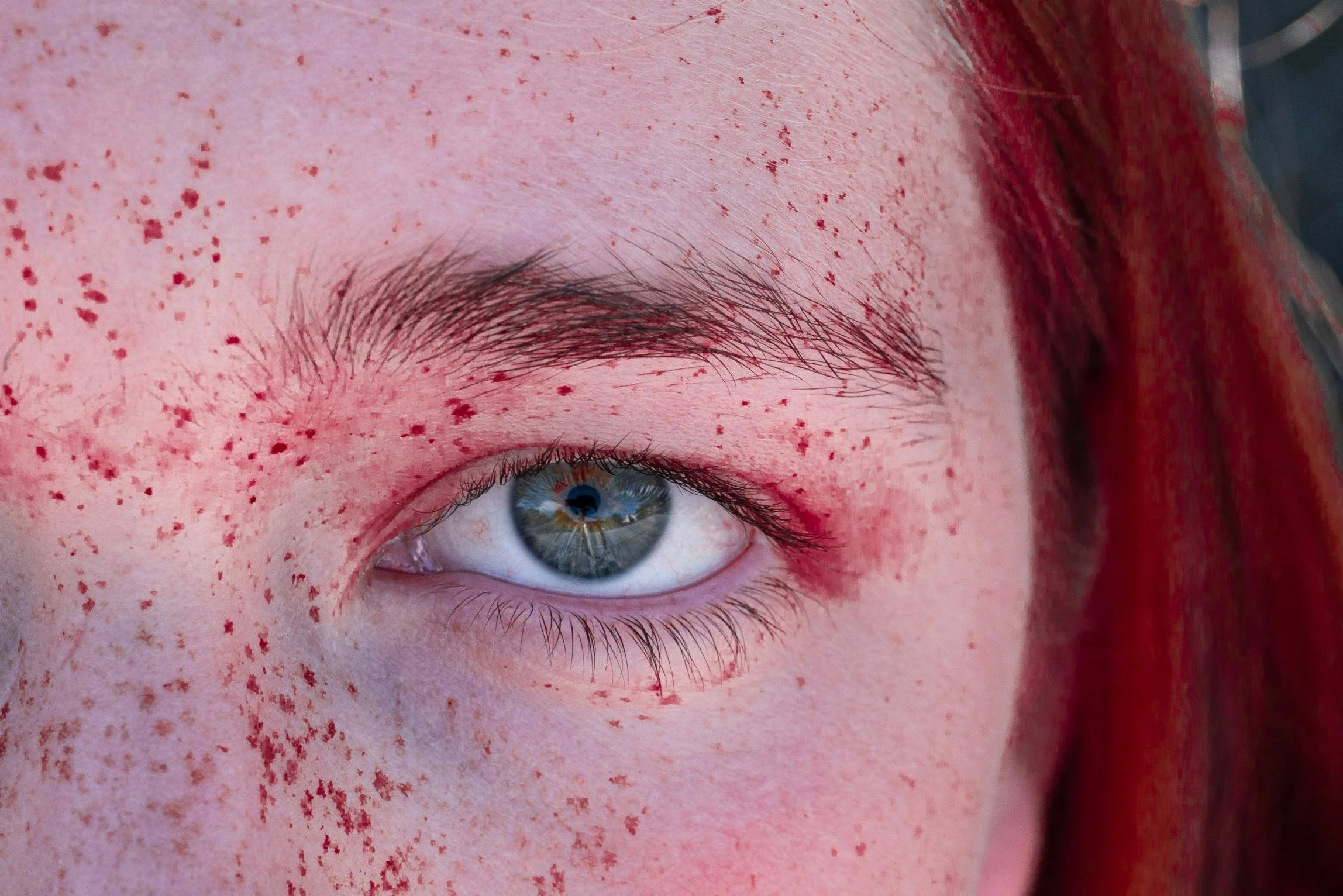 Close-up of a person's eye with light-colored iris, surrounded by freckles on fair skin, with red hair visible on the right side.