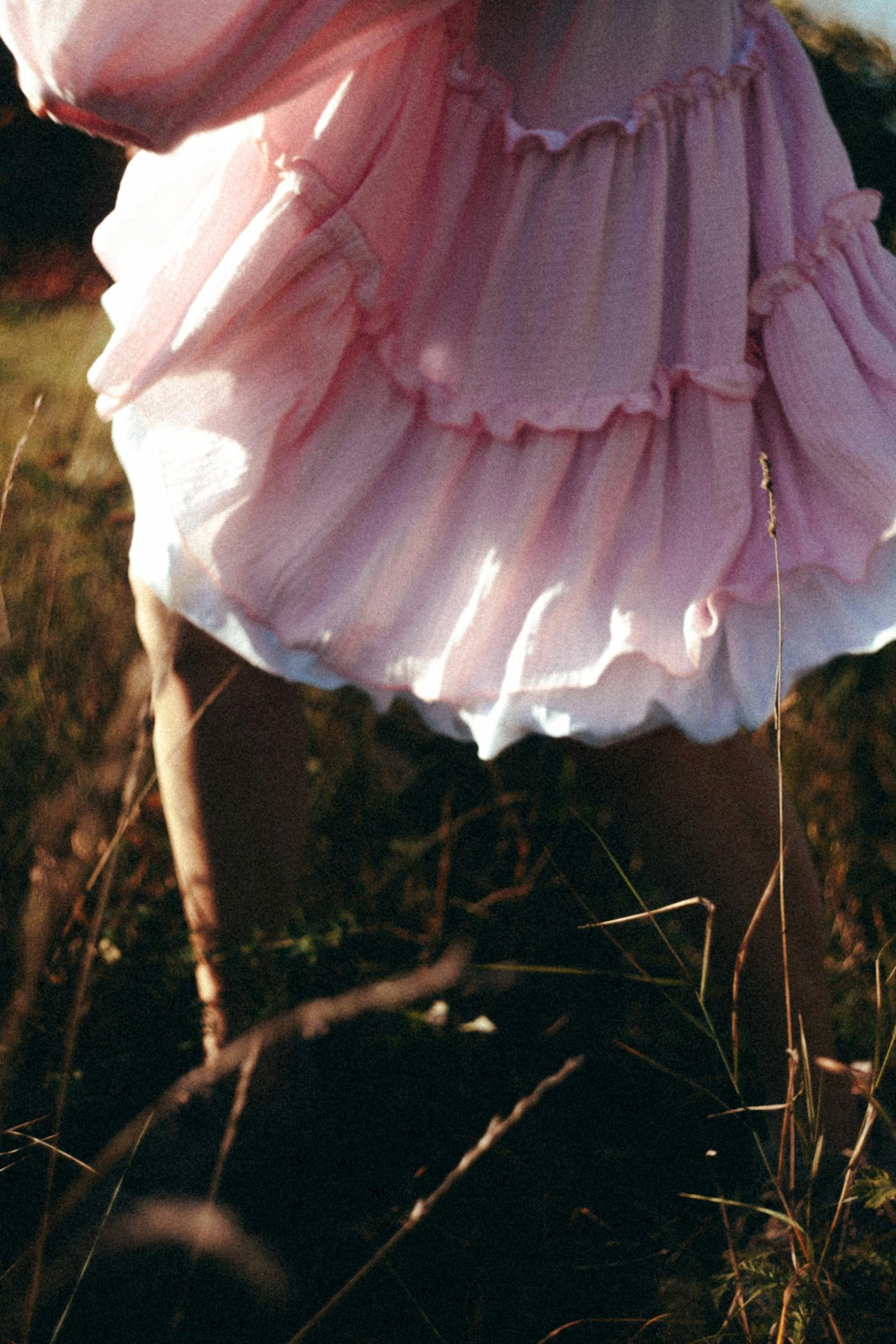 A person wearing a pink ruffled dress standing outdoors on grass and small plants.