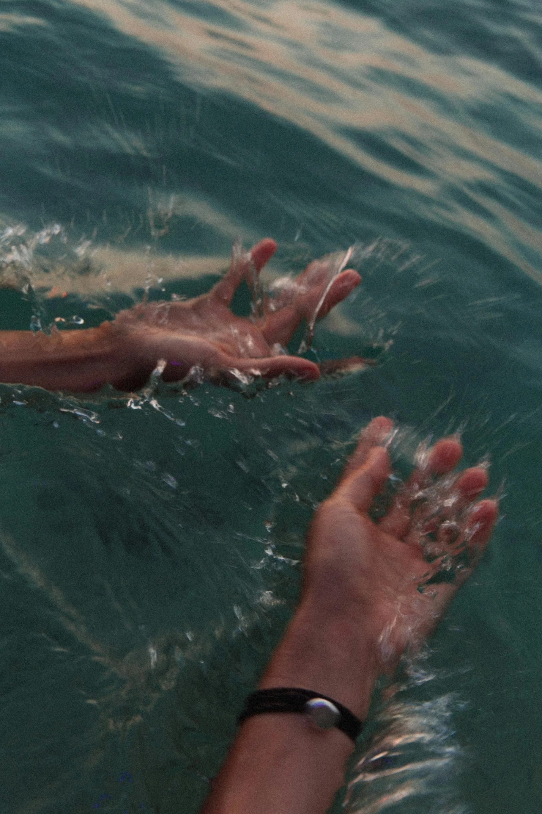 Two hands reaching into the water, one with a bracelet, as a third hand is submerged, creating ripples and splashes.