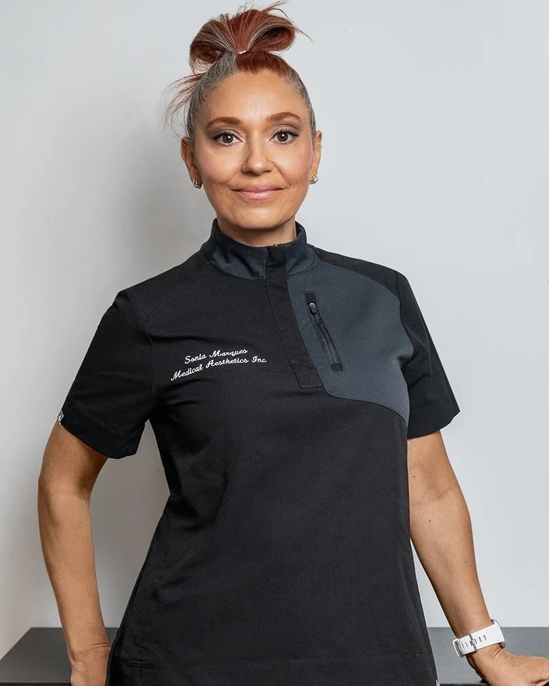 A woman with light skin, styled in a high bun and wearing small hoop earrings, is standing against a plain background. She is dressed in a dark-colored, short-sleeved medical uniform with a zippered collar and a name tag that reads 'Sonia Marqueso Medical Aesthetics Inc.' She has a subtle smile, and her right hand is resting on a surface while her left arm is relaxed by her side with a white wristwatch.