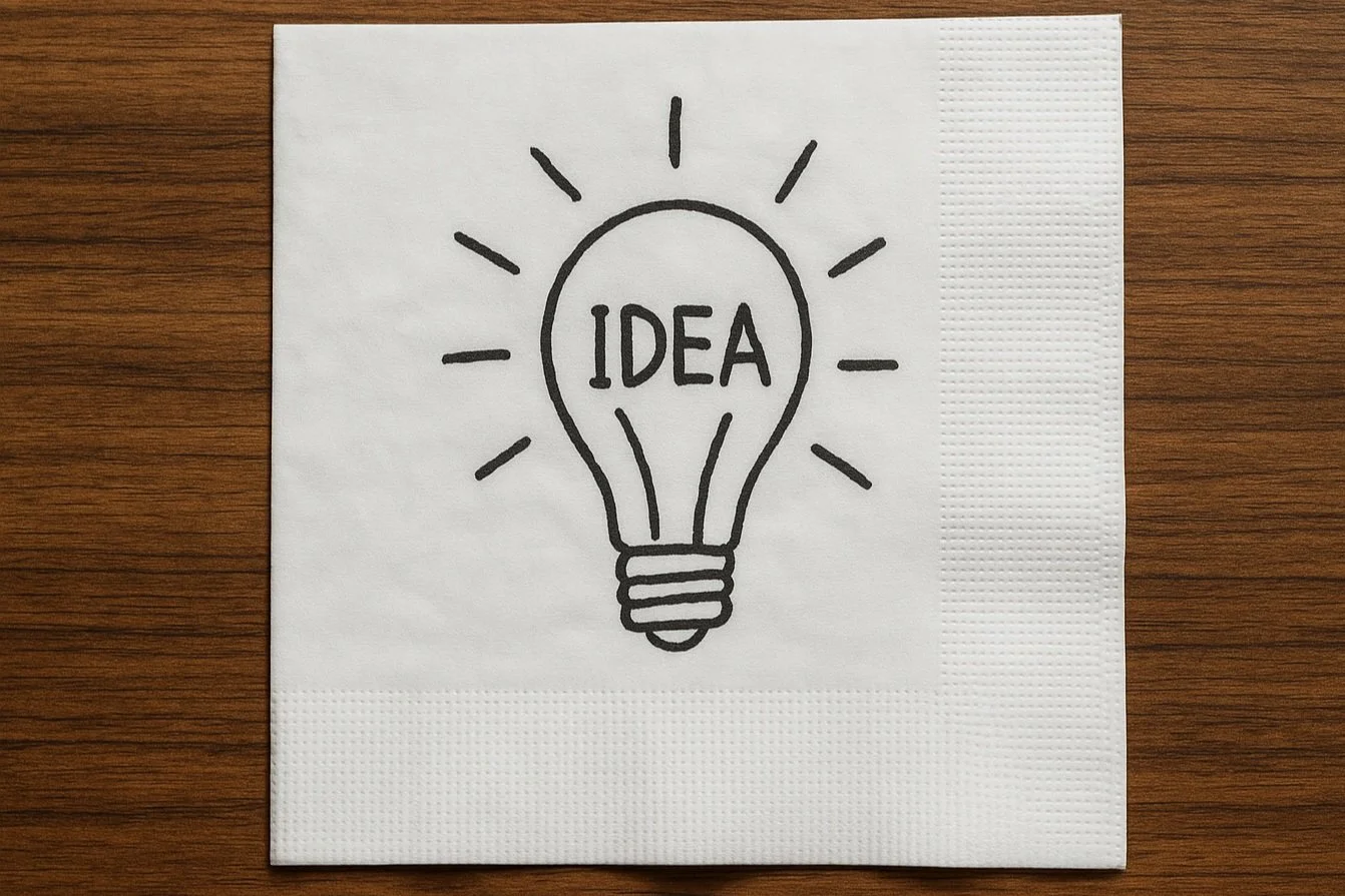 A paper napkin with a black line drawing of a light bulb and the word 'IDEA' inside, placed on a wooden surface.