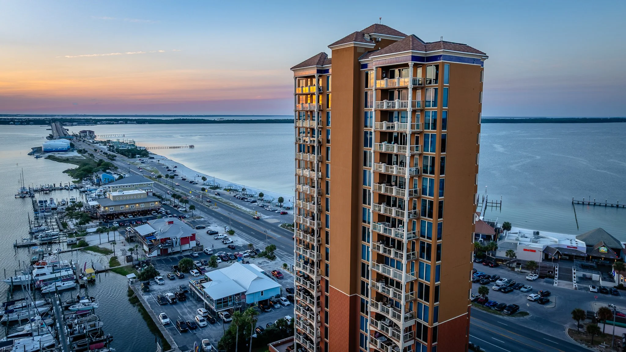 Drone photo of hotel near a body of water with boats, marina, and parking lots at sunset in Pensacola Beach