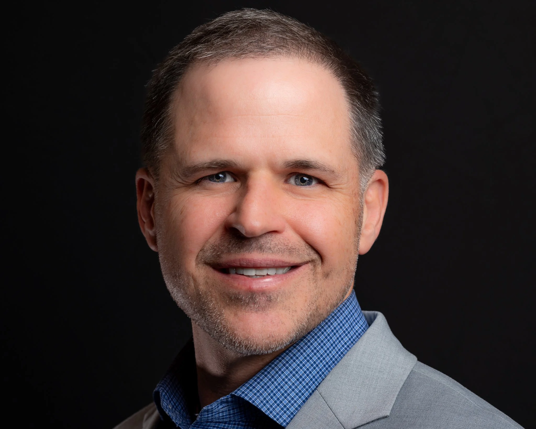 Headshot of a smiling professional with short hair, blue eyes, wearing a blue checkered shirt and light gray blazer against a black background.