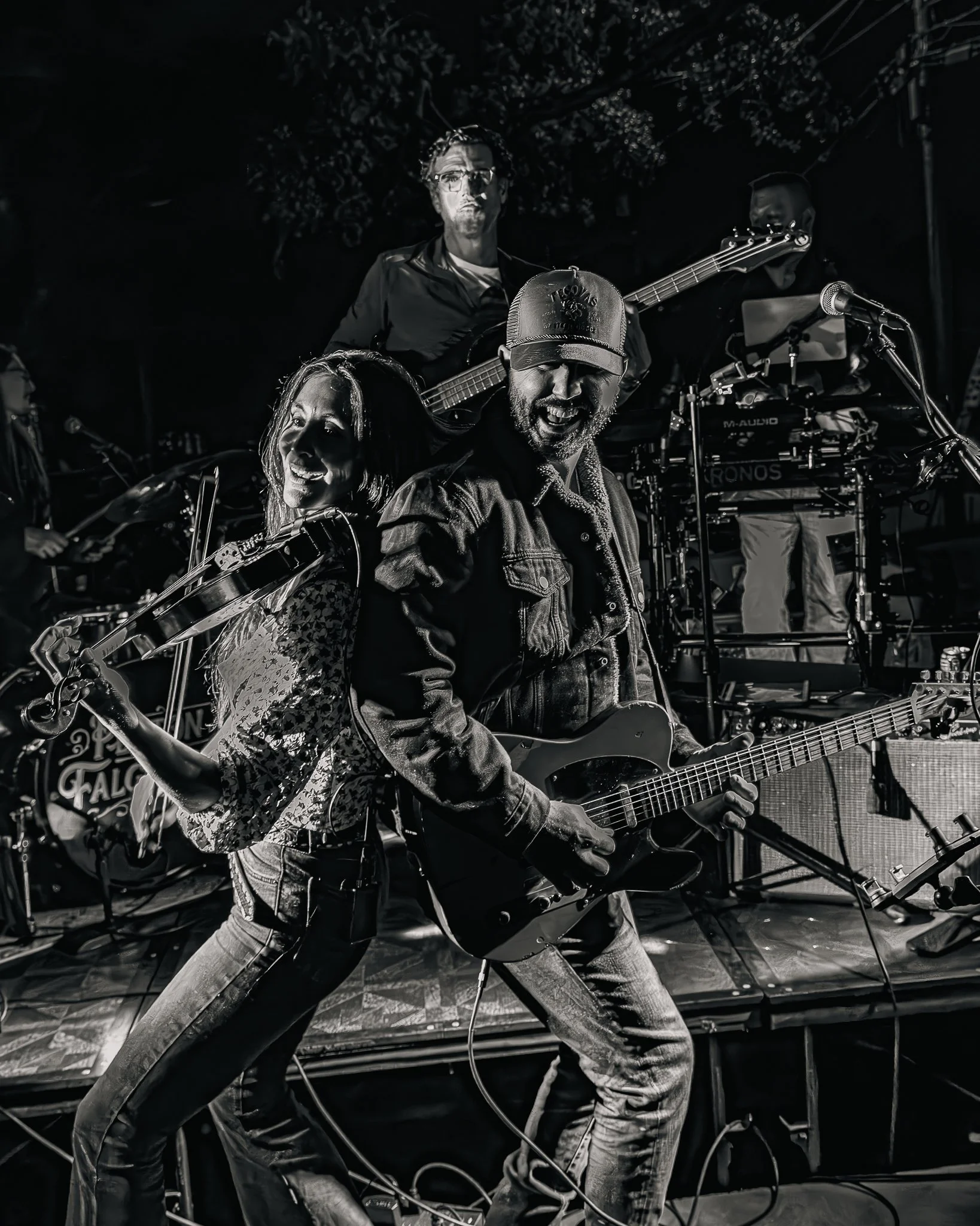 A live band performing on stage, with a guitarist, a violinist, a bass player, and a keyboard player, all passionately engaged in their music in Mid City, Baton Rouge