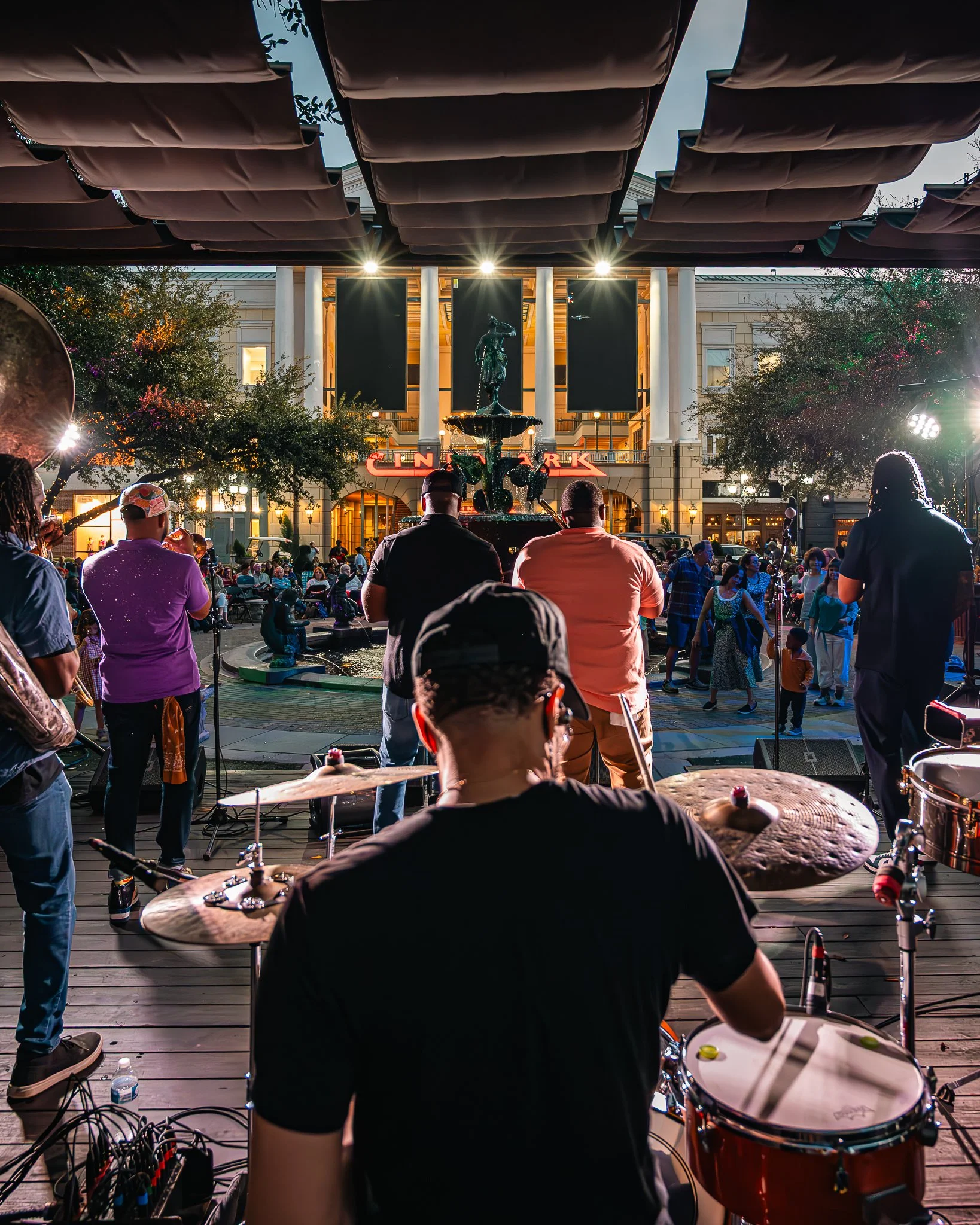 A live outdoor music performance at night in Perkins Rowe with a band playing, audience dancing, and a fountain in the center. The scene is lit by stage lights and surrounding buildings. Located in Baton Rouge