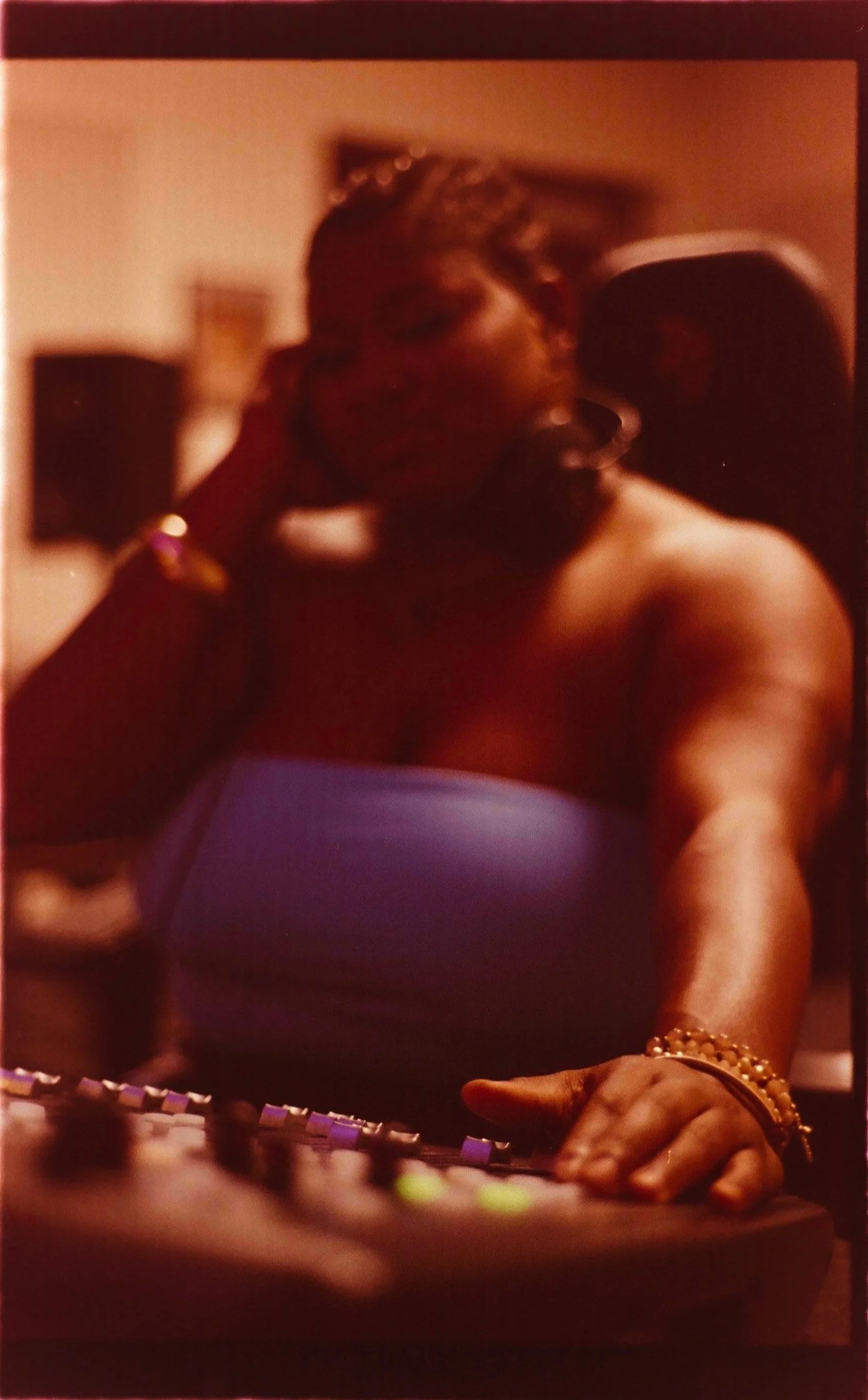 A woman in a blue strapless top, wearing jewelry, is in a recording studio with a microphone in front of her, possibly singing or speaking into it. She is touching a soundboard or mixing console.