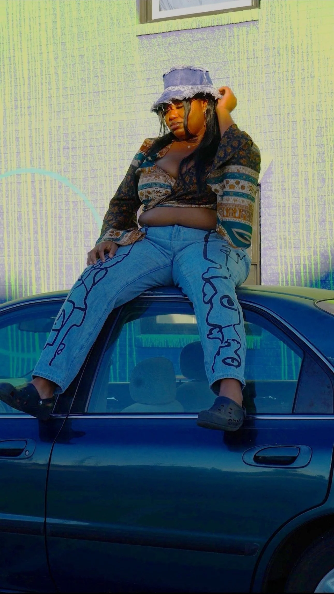 A person sitting on the roof of a dark green car, wearing a bucket hat, a patterned blouse, and jeans with abstract designs. The person is touching their hat and appears to be looking down or to the side. The background features a house with a textured wall and a window.