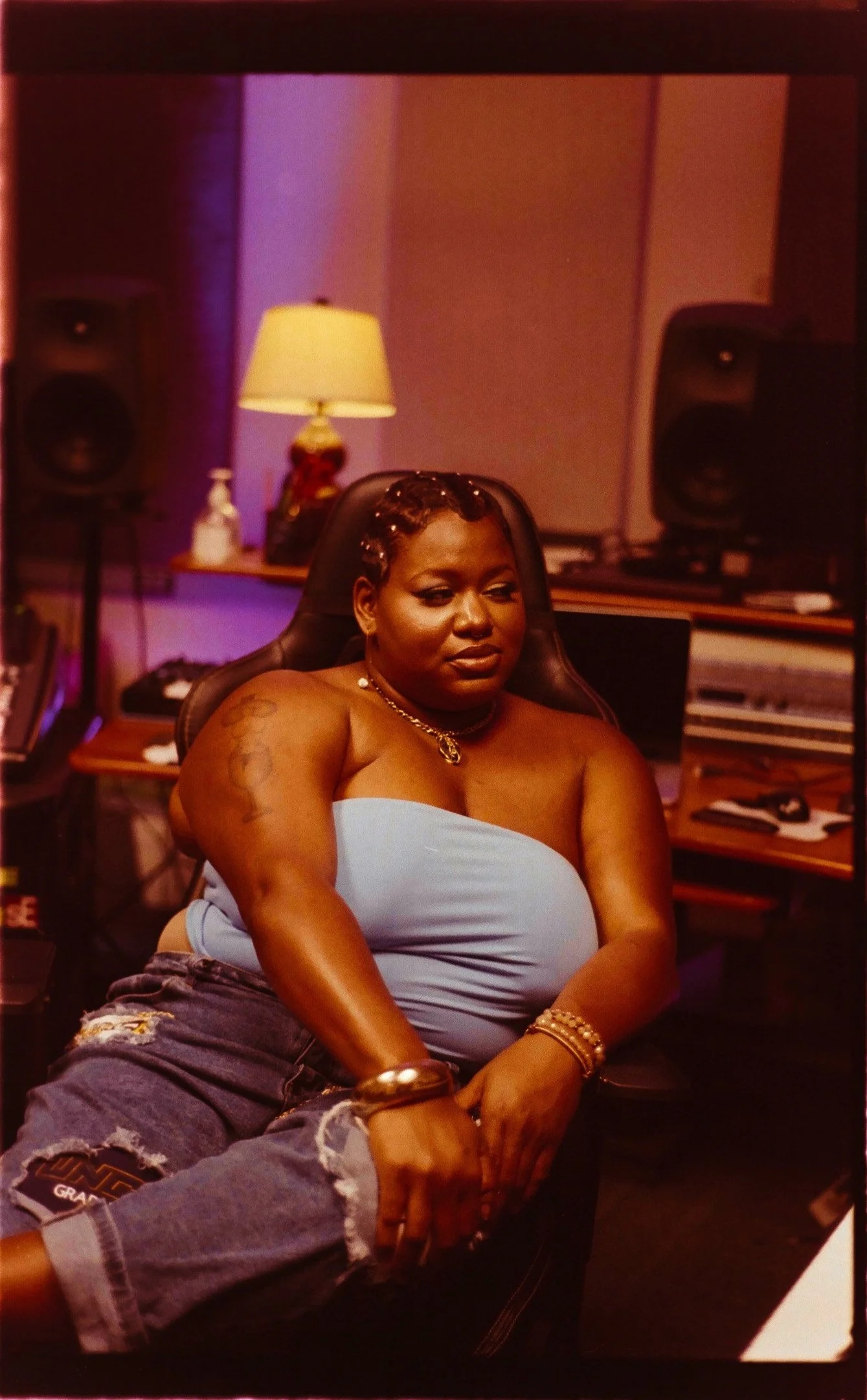 A woman with dark skin, wearing a light blue strapless top and ripped denim shorts, sitting in a recording studio surrounded by musical equipment and a lamp, with a calm expression.