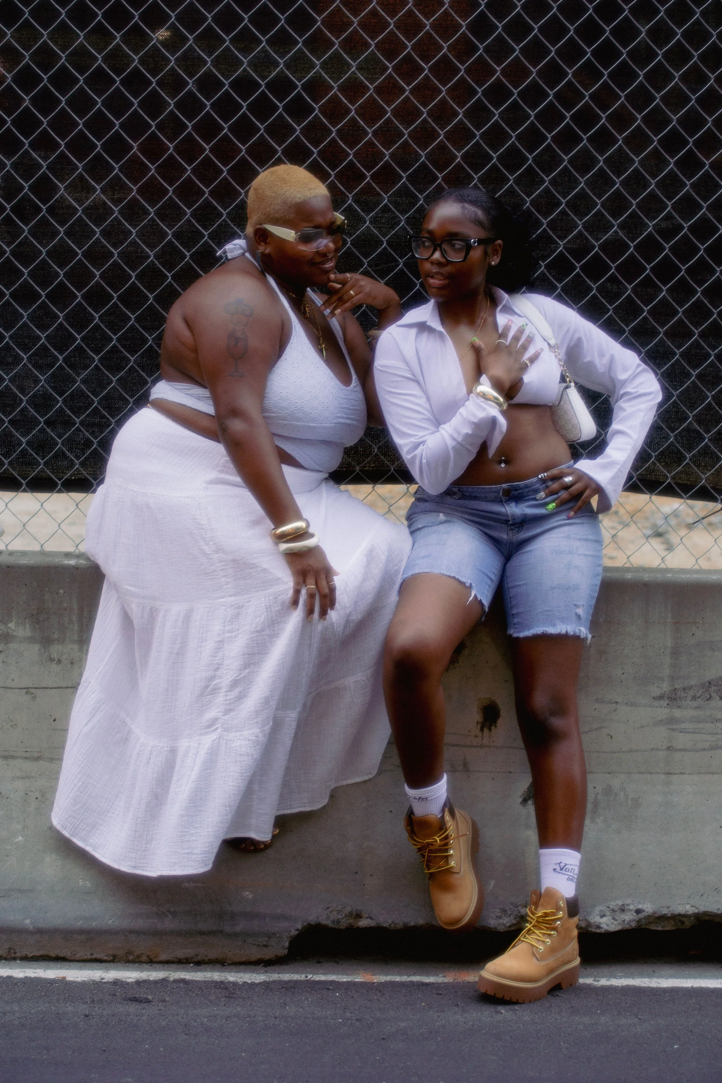 Two women talking near a chain-link fence, one wearing a white dress and the other in a white shirt, denim shorts, and tan boots.