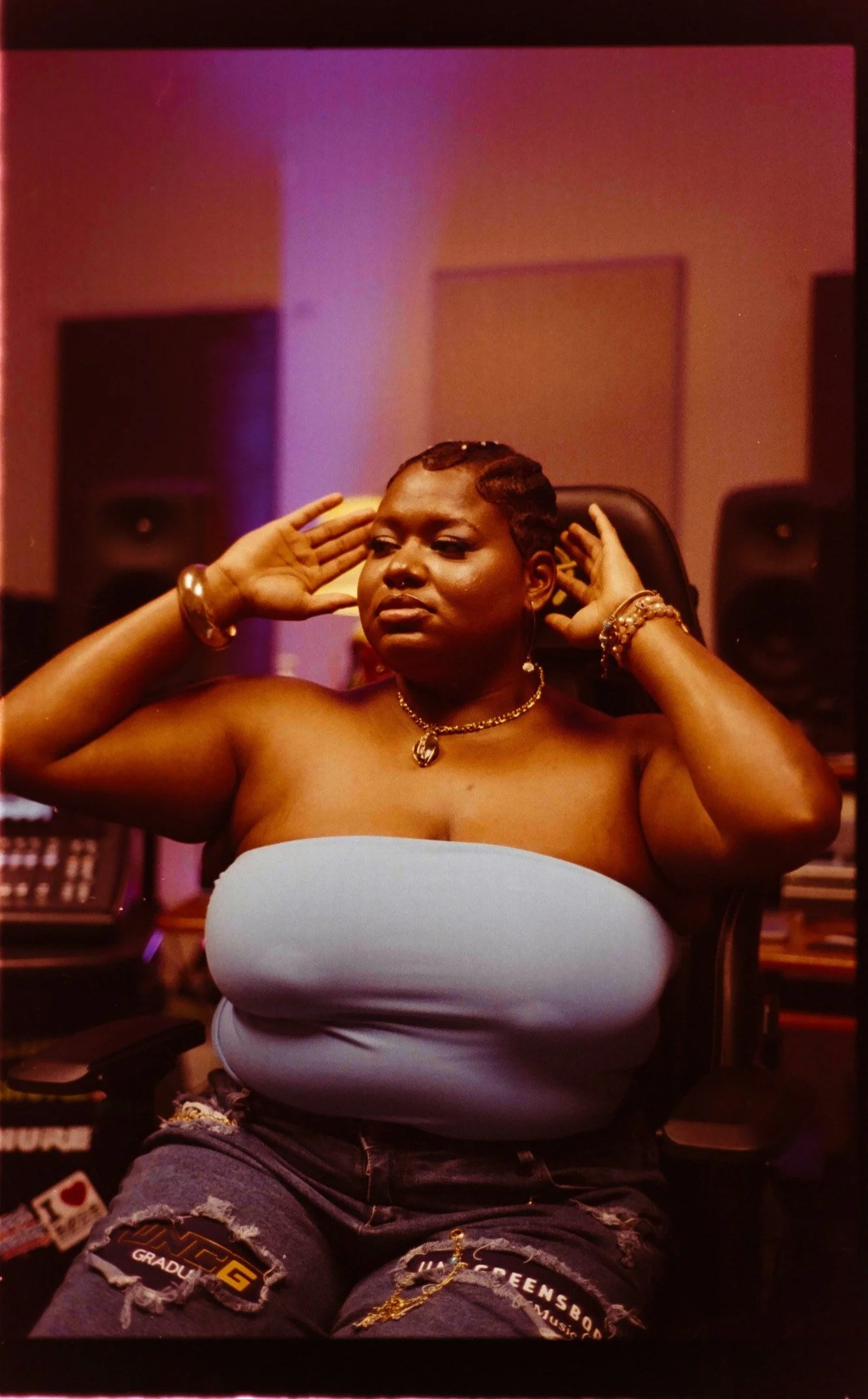 A woman with short hair and jewelry is sitting in a recording studio, adjusting her headphones with her eyes closed, wearing a strapless top and ripped jeans with patches.