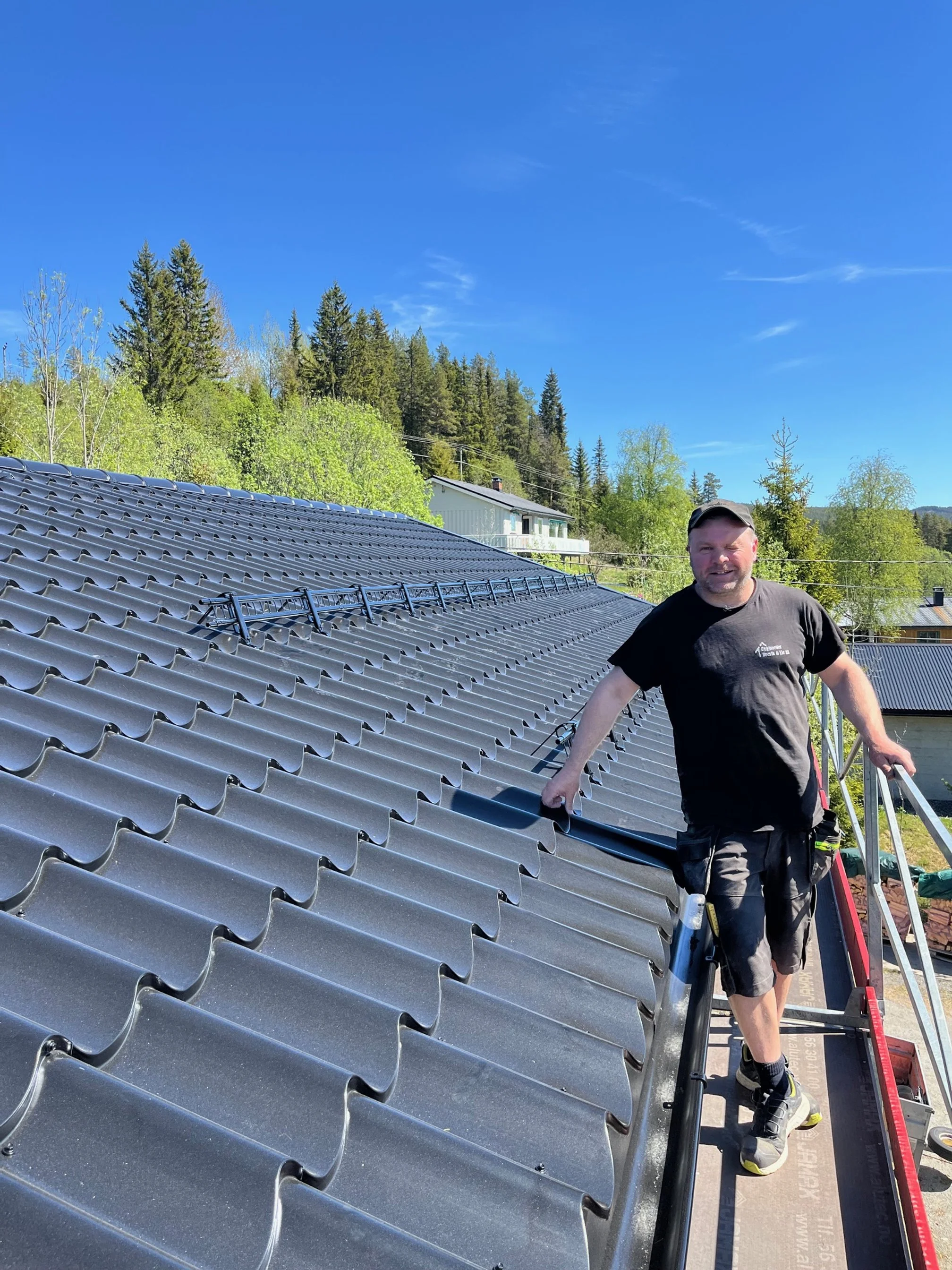 A man installing solar panels on a metal roof in a residential area with green trees and houses in the background on a sunny day.